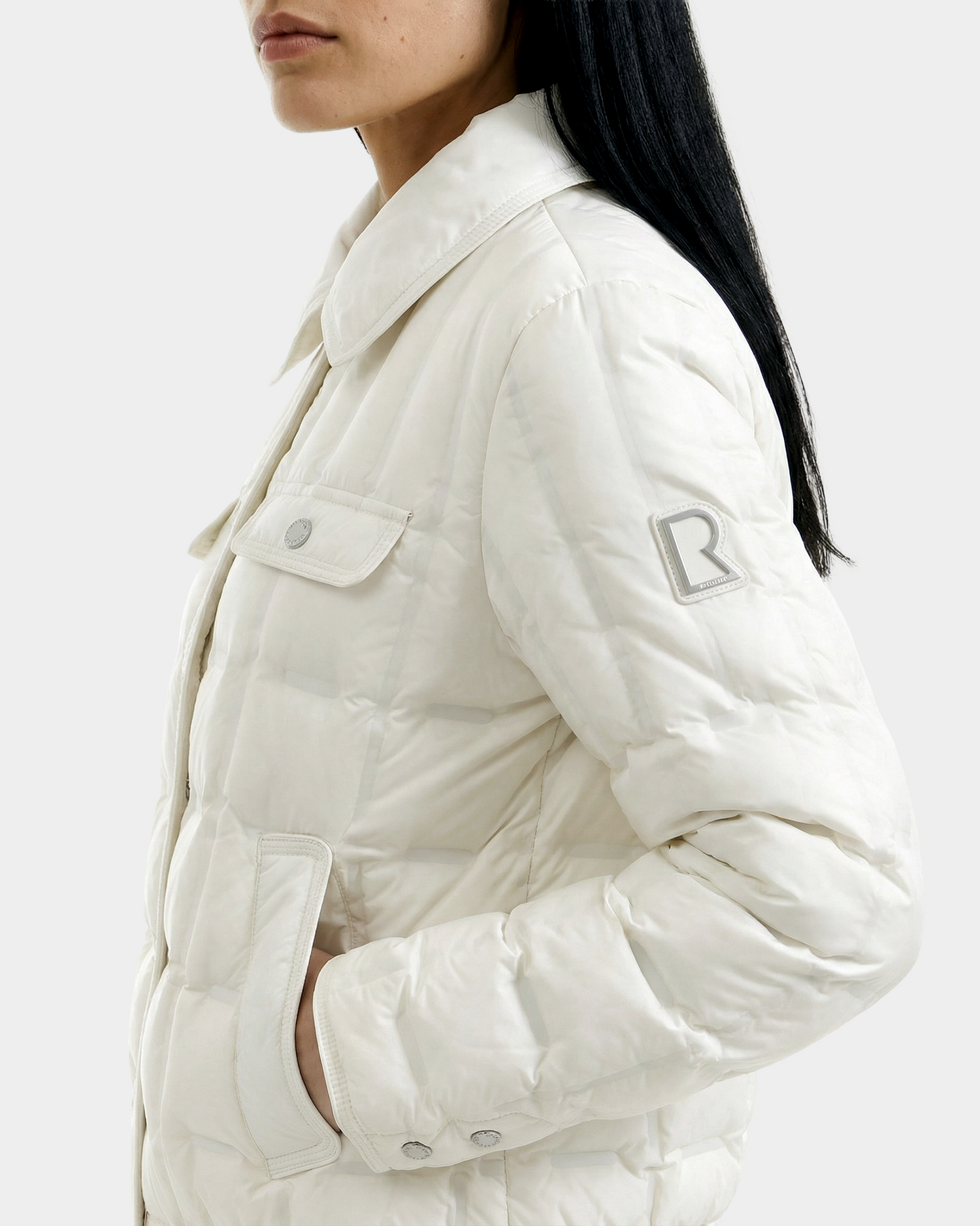 A woman with long dark hair wears the Rudsak SEOUL - 8126027 Iceland quilted down shirt jacket, featuring lightweight warmth, a chest pocket, and an R logo on the sleeve. Only her lower face is visible, and her hand rests in the pocket.