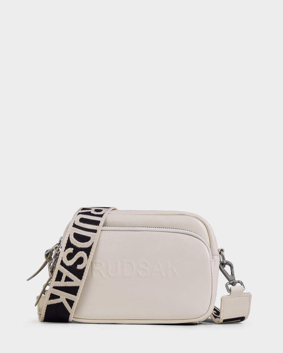 The ZOYA L - 8325543 Vanilla by Rudsak is a small rectangular white leather crossbody bag with RUDSAK embossed on the front, an adjustable black-and-white logo strap, silver hardware, and a front zipper pocket for a stylish finish.
