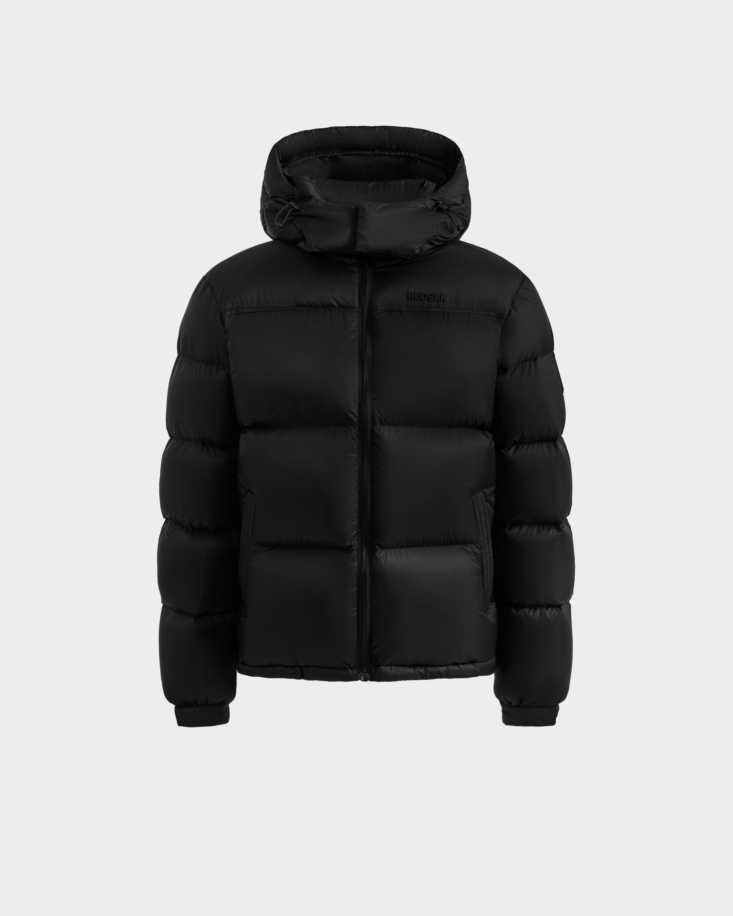 The STEFANO - 6125574 Black by Rudsak is a black men’s down puffer jacket with a hood, front zipper, and quilted sections, shown against a light gray background—a sleek technical outerwear choice.
