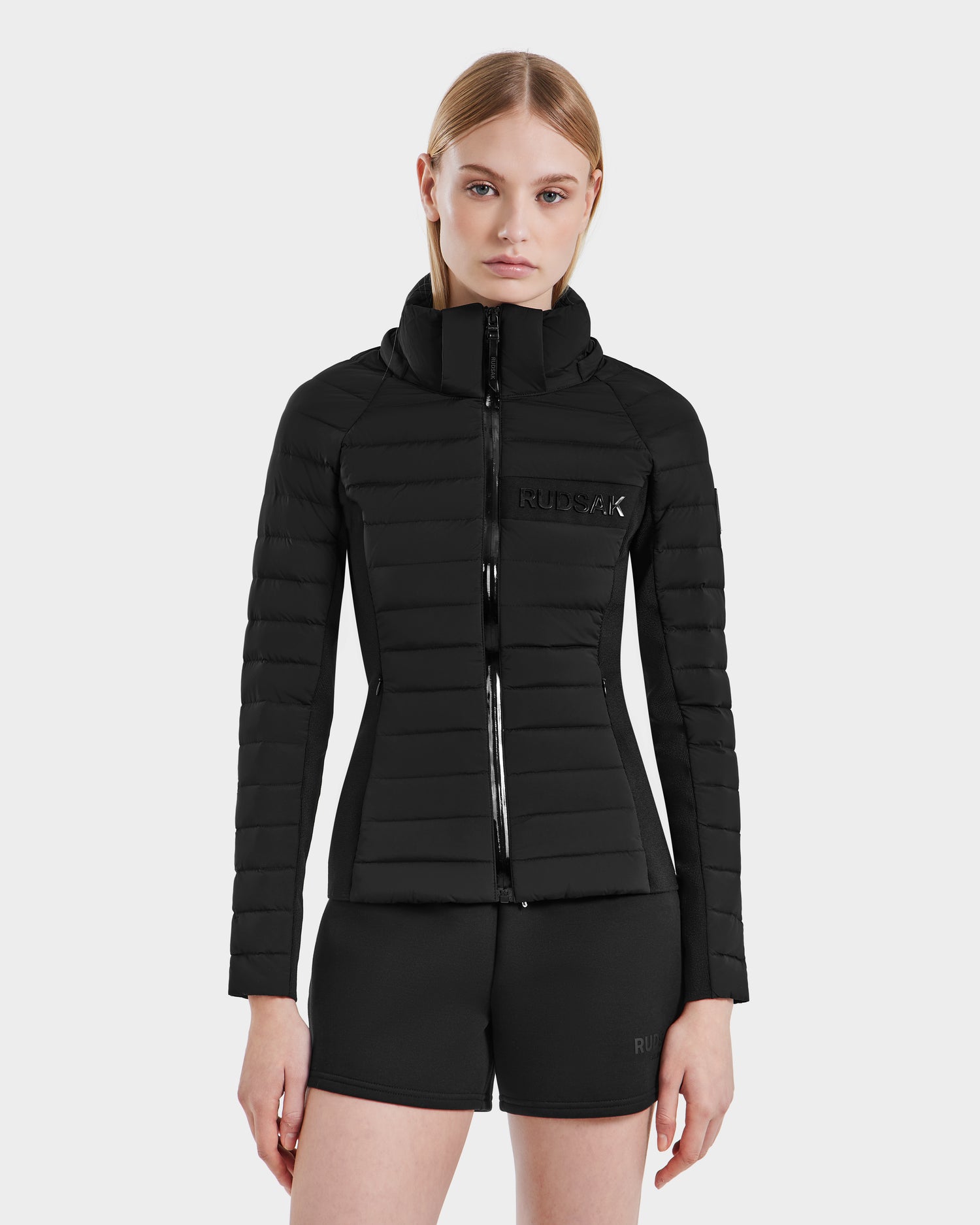 A woman with straight blonde hair wears the RUDSAK ALANA - 8123022 BLACK fitted women's down jacket with a hood and black shorts, standing facing forward against a plain white background.