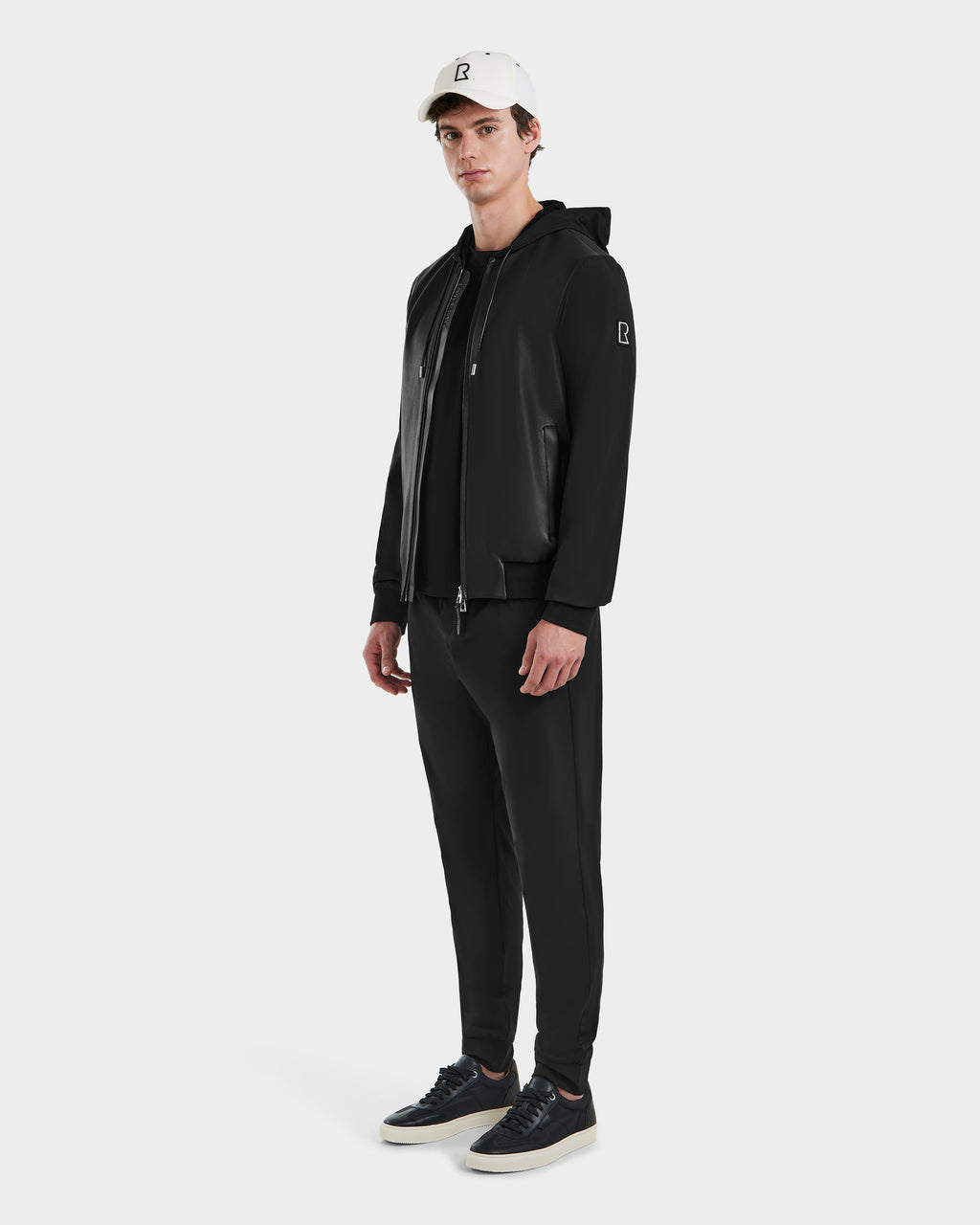 A person stands facing forward against a plain white background, wearing the Rudsak ALBY - 6125033 Black bomber jacket, paired with black jogger pants, black sneakers, and a white baseball cap featuring a small black logo.