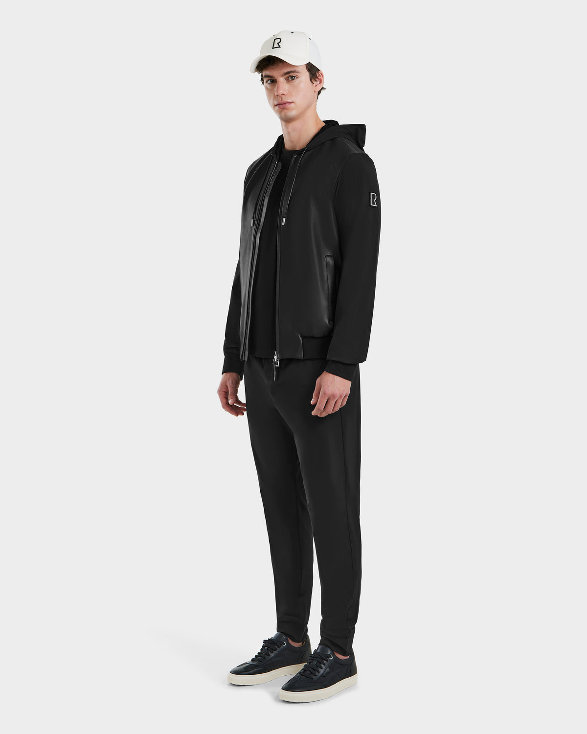 A person stands facing forward against a plain white background, wearing the Rudsak ALBY - 6125033 Black bomber jacket, paired with black jogger pants, black sneakers, and a white baseball cap featuring a small black logo.