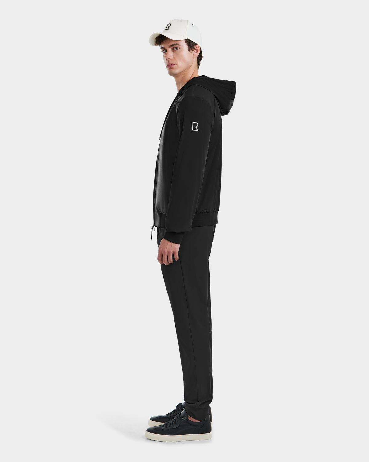 A person in profile view faces left wearing Rudsaks ALBY - 6125033 Black outerwear, black pants, black sneakers, and a white baseball cap against a plain light background.