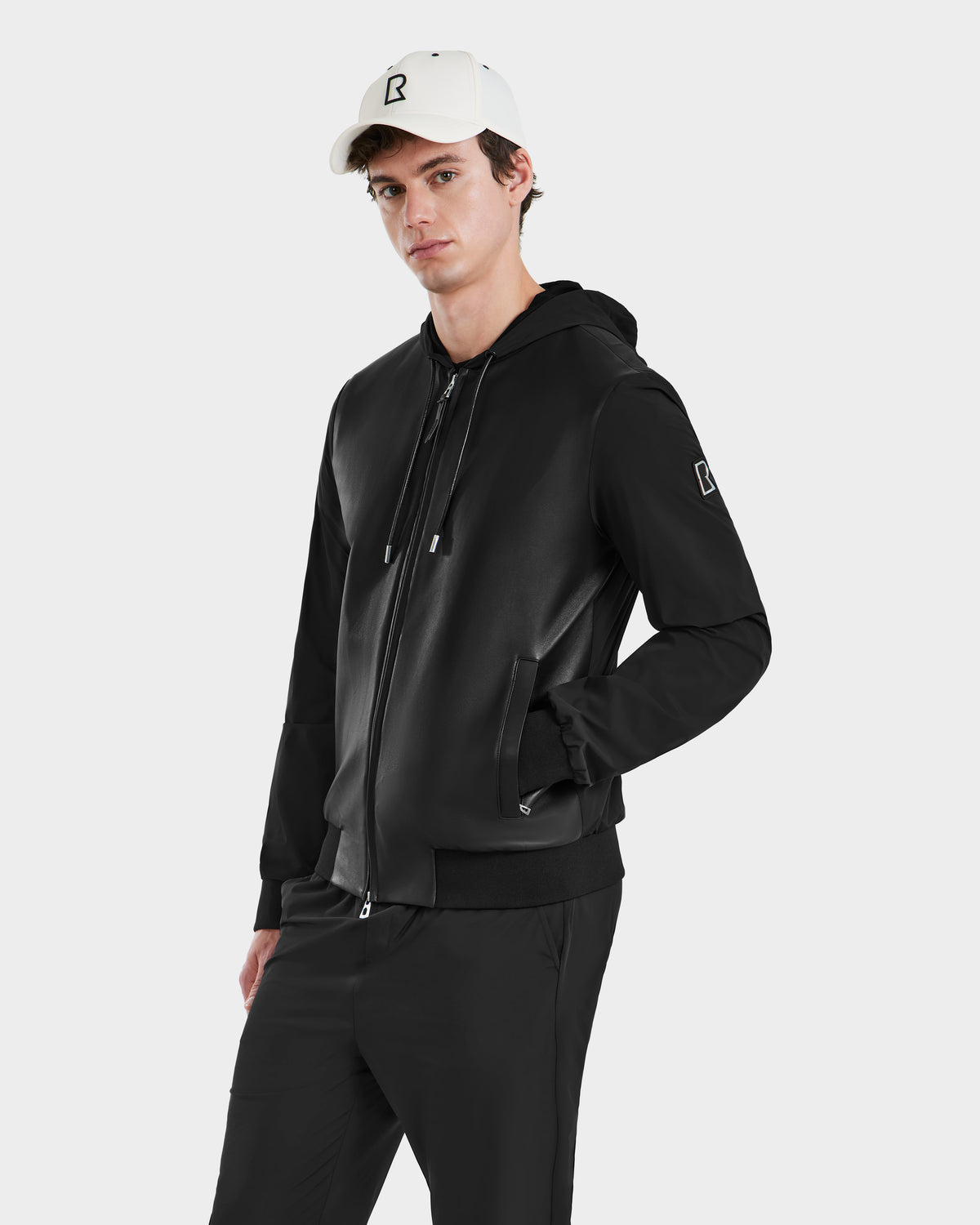 A young man models the Rudsak ALBY - 6125033 Black bomber jacket paired with black pants and a white cap, standing against a plain white background to showcase this versatile outerwear for any occasion.