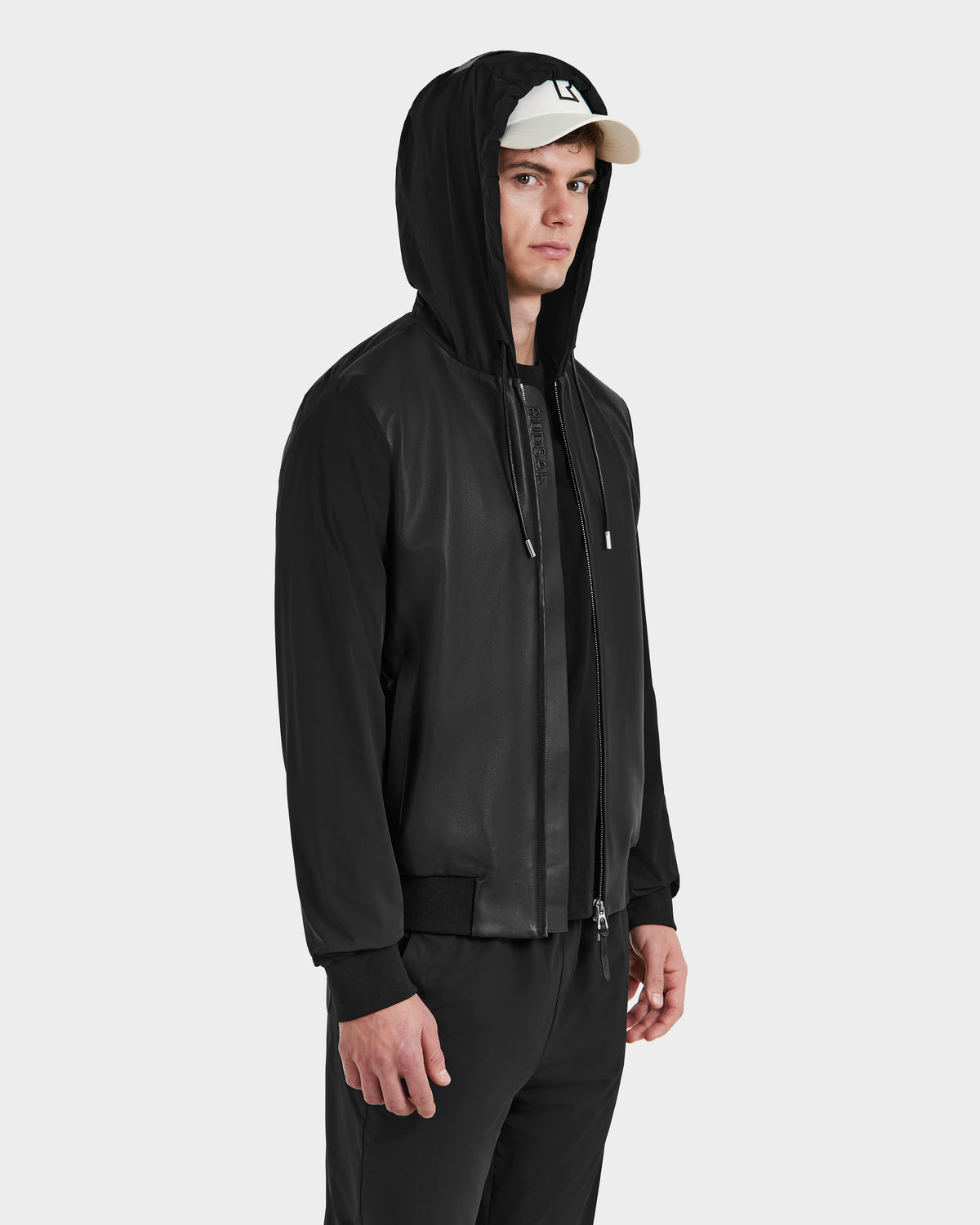 A man wearing the Rudsak ALBY - 6125033 Black bomber jacket with a leather front, paired with a cap.