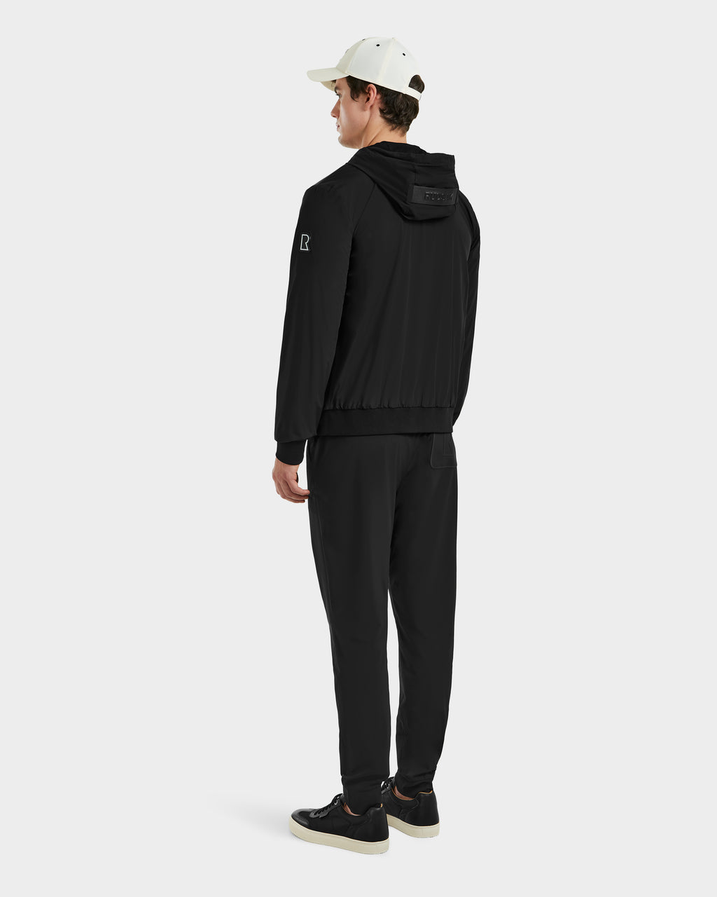 A man faces away in a Rudsak ALBY - 6125033 Black bomber jacket, black pants, white-capped and wearing black sneakers with white soles against a light background—highlighting versatile outerwear for any casual look.