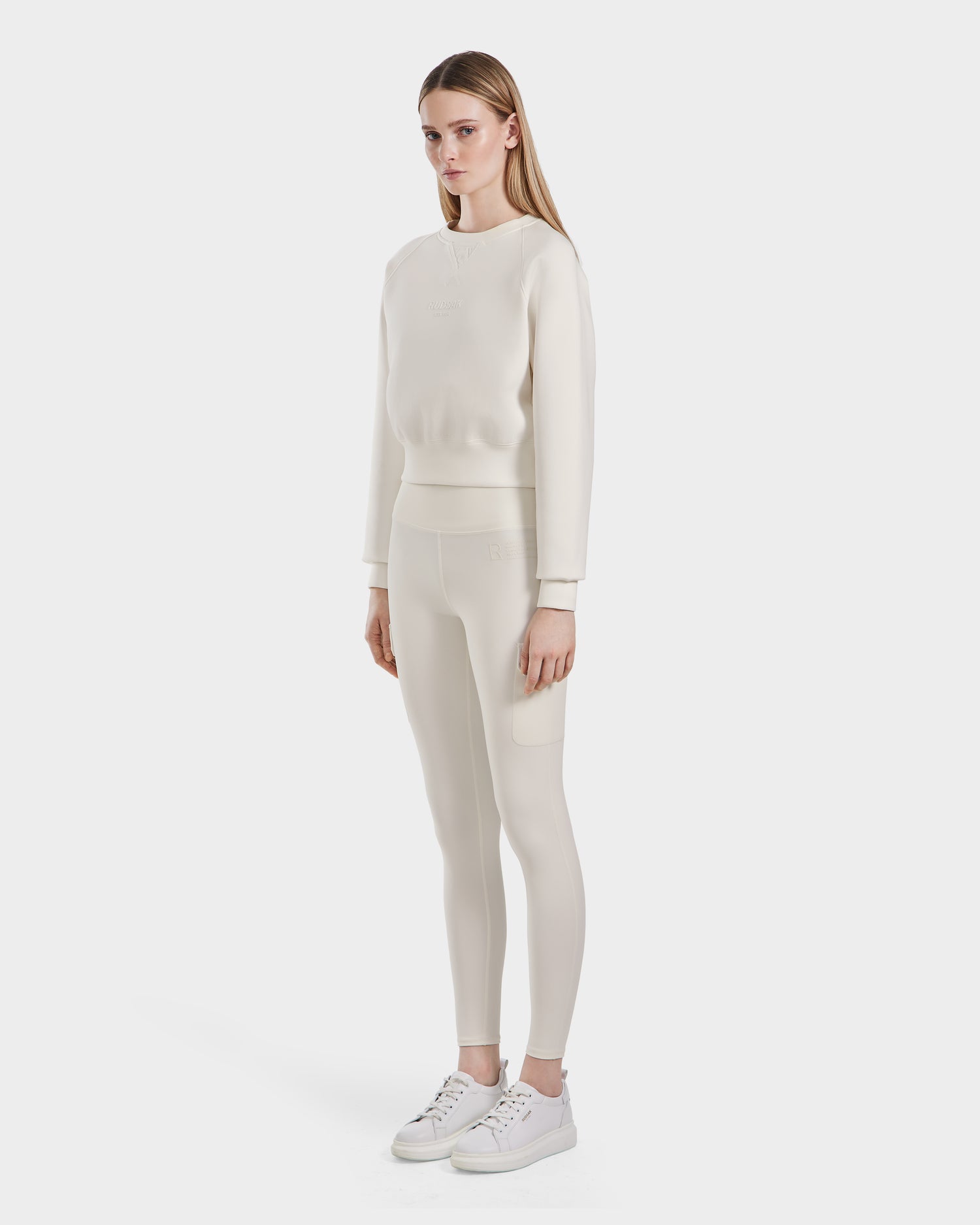 A woman faces forward, wearing the Rudsak BETSY - 8525087 Iceland matching cream premium cotton blend crewneck sweater and leggings set with white sneakers, against a plain white background.