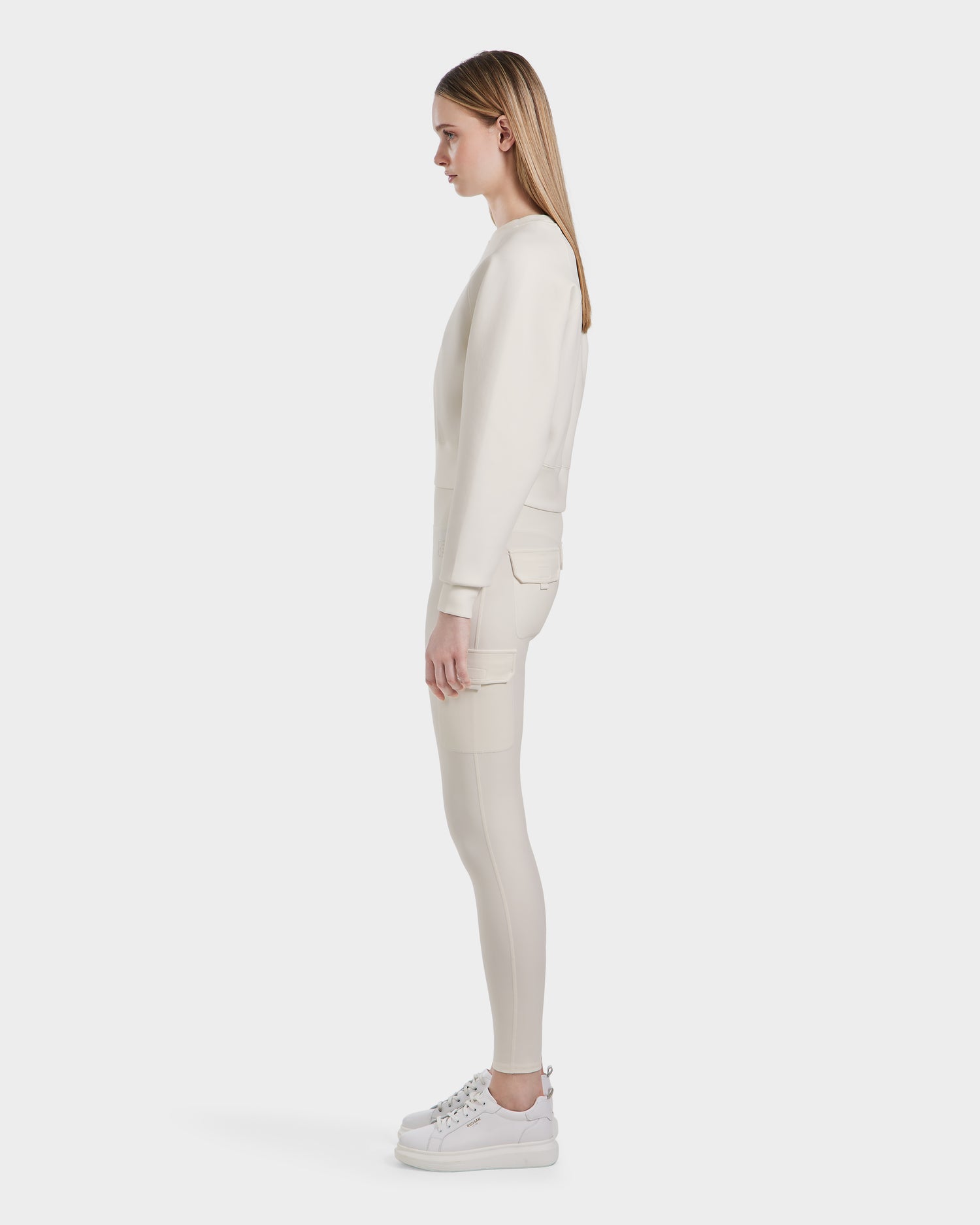 A woman with straight, light brown hair stands in profile wearing the Rudsak BETSY - 8525087 Iceland premium cotton-blend women's crop sweater and matching off-white leggings with sneakers against a plain white background.