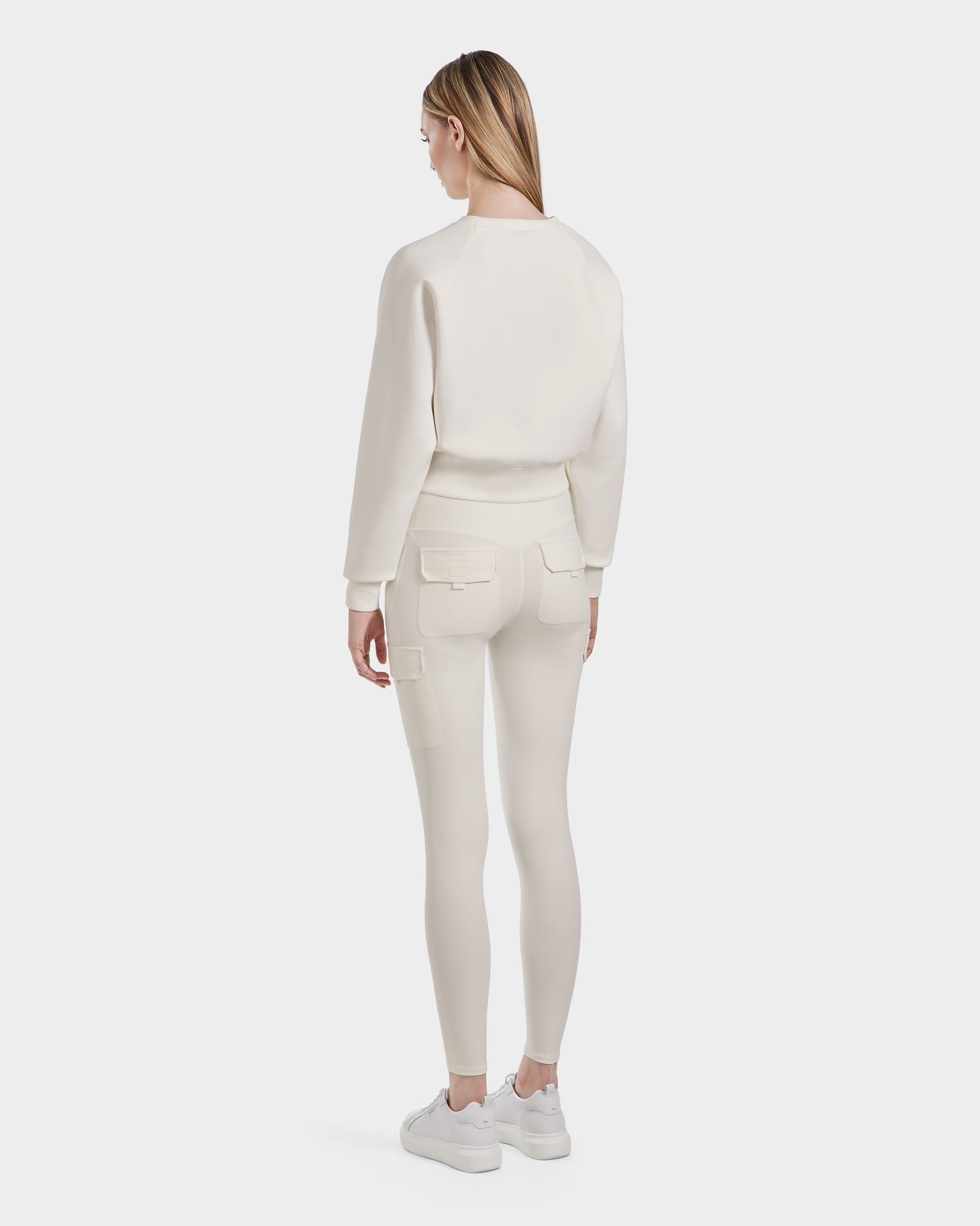 A woman with straight, light brown hair faces away, showcasing the Rudsak BETSY - 8525087 Iceland off-white crewneck sweater and matching fitted pants with flap pockets, paired with white sneakers against a plain white background.