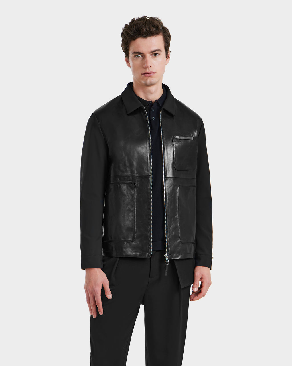 A man with short brown hair stands against a light gray background, wearing the Rudsak BRYAN - 6125016 Black shirt jacket over a black shirt and black pants, highlighting versatile men’s outerwear.