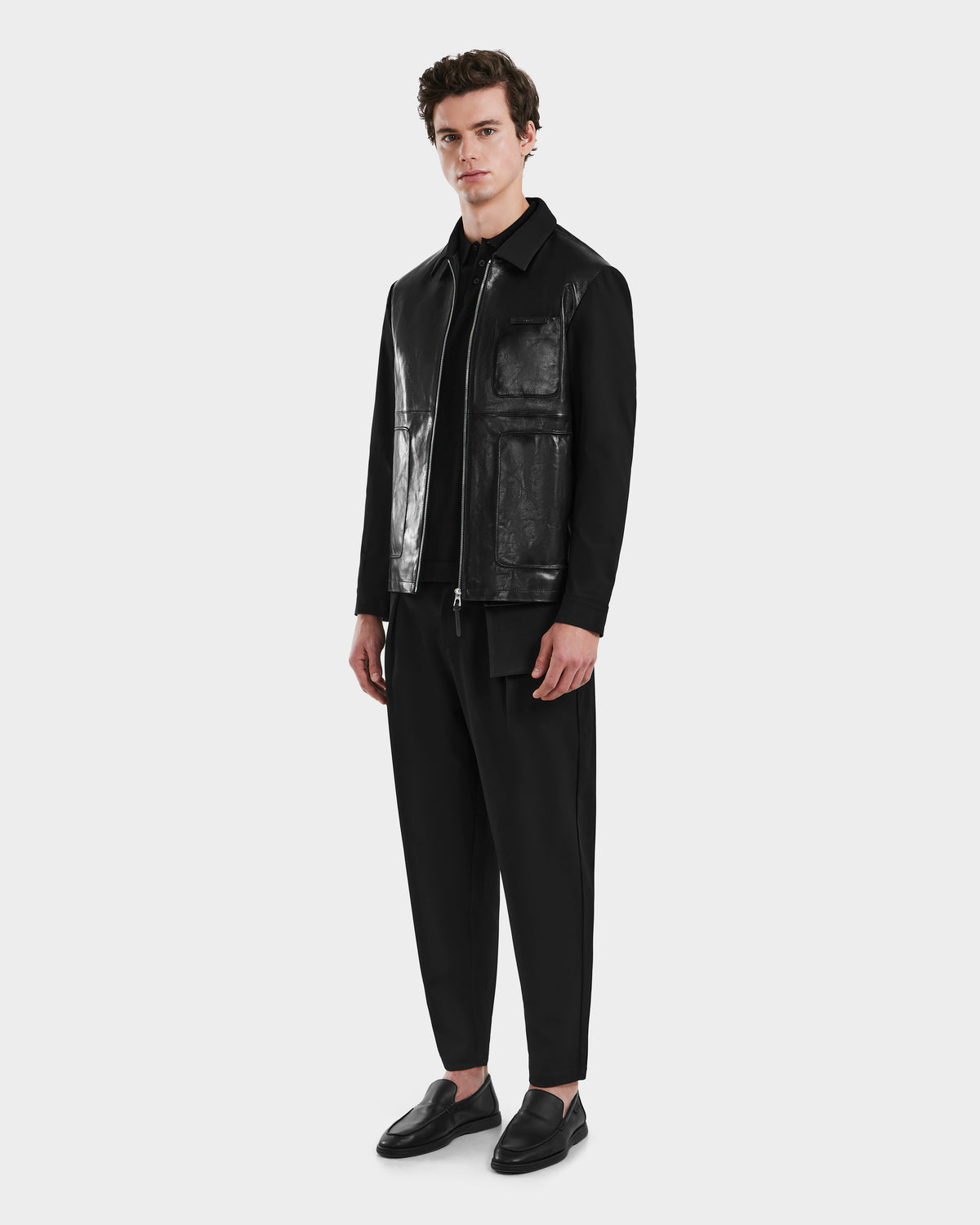 A man with short brown hair models the Rudsak BRYAN - 6125016 Black glossy leather shirt jacket against a plain background, styled with black pants and loafers for a versatile outerwear look.