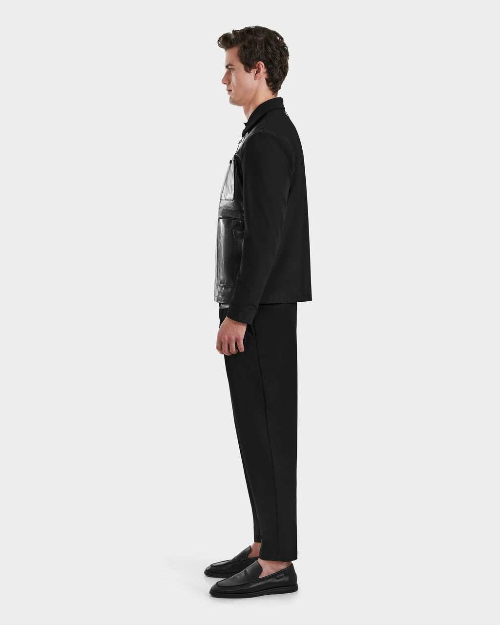 A young man stands in profile facing left, wearing Rudsak’s BRYAN - 6125016 Black shirt jacket, black pants, and black shoes against a plain white background.