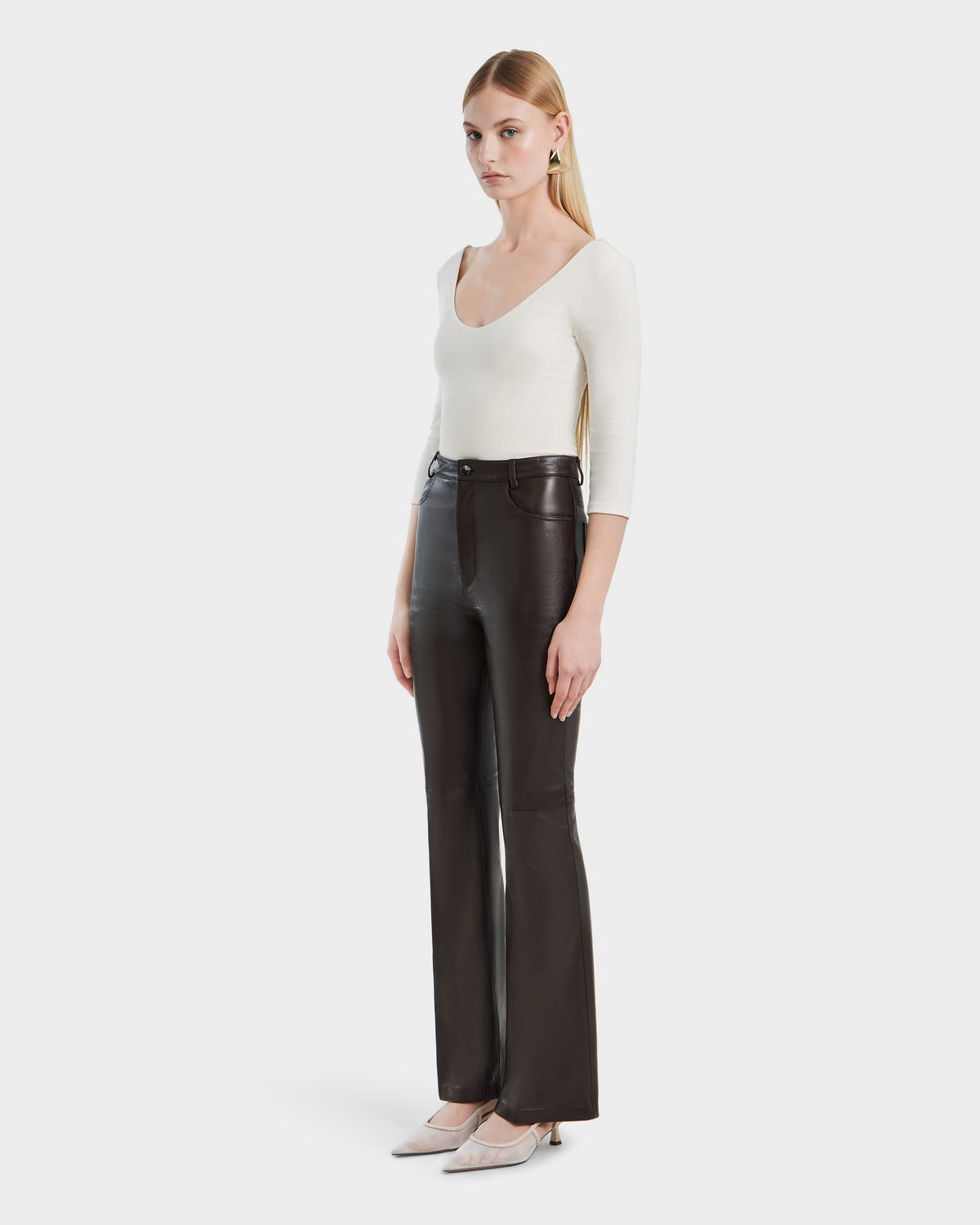 A woman with long blonde hair wears a fitted white long-sleeve top, light-colored pointed-toe heels, and Rudsak CARISSA - 8524545 Espresso leather pants, standing against a plain white background.
