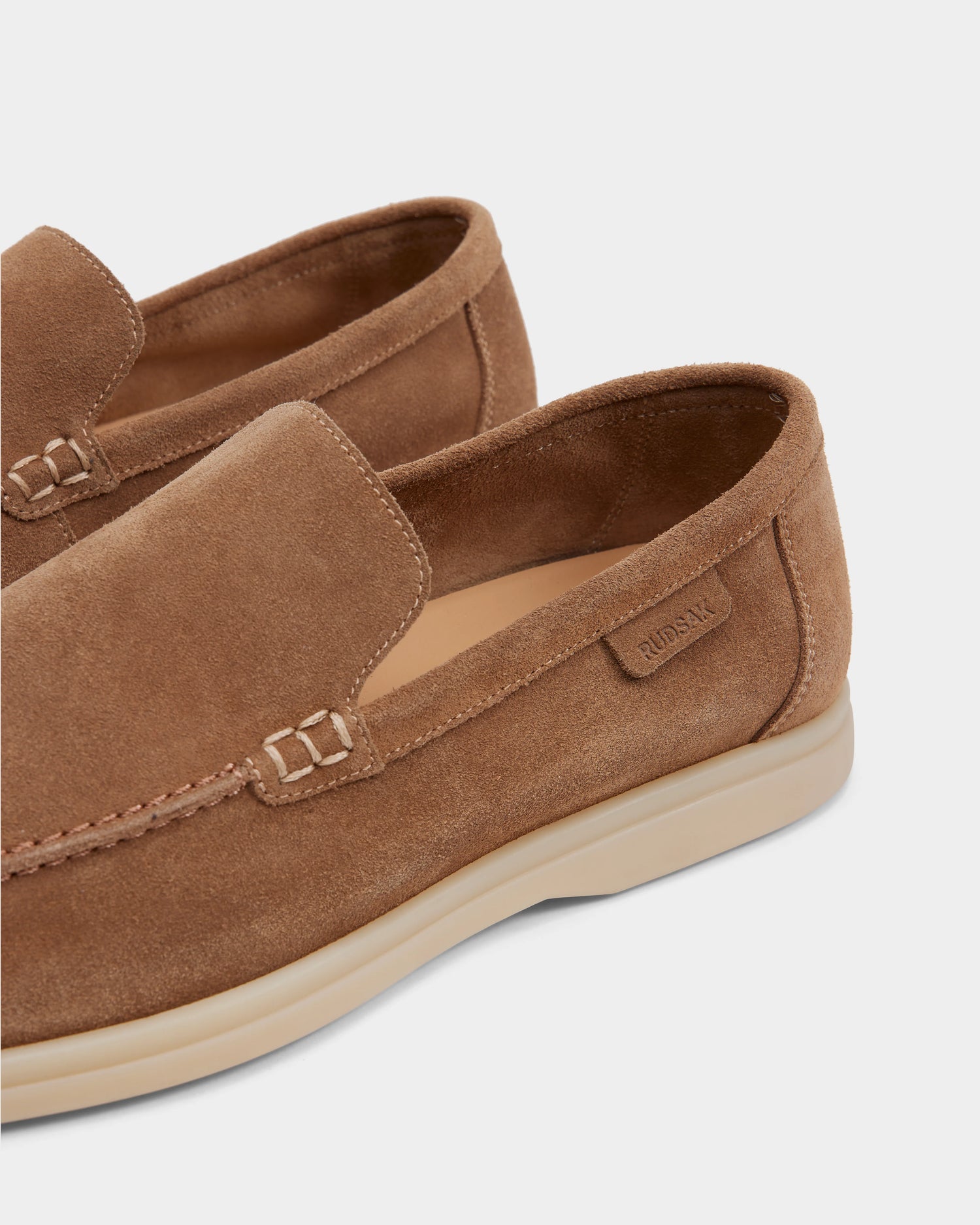 Close-up of premium men’s footwear: Rudsak CHANNING - 6225005 Almond suede slip-on loafers in light brown, featuring light beige rubber soles, detailed stitching, and a small branded tag on the side.