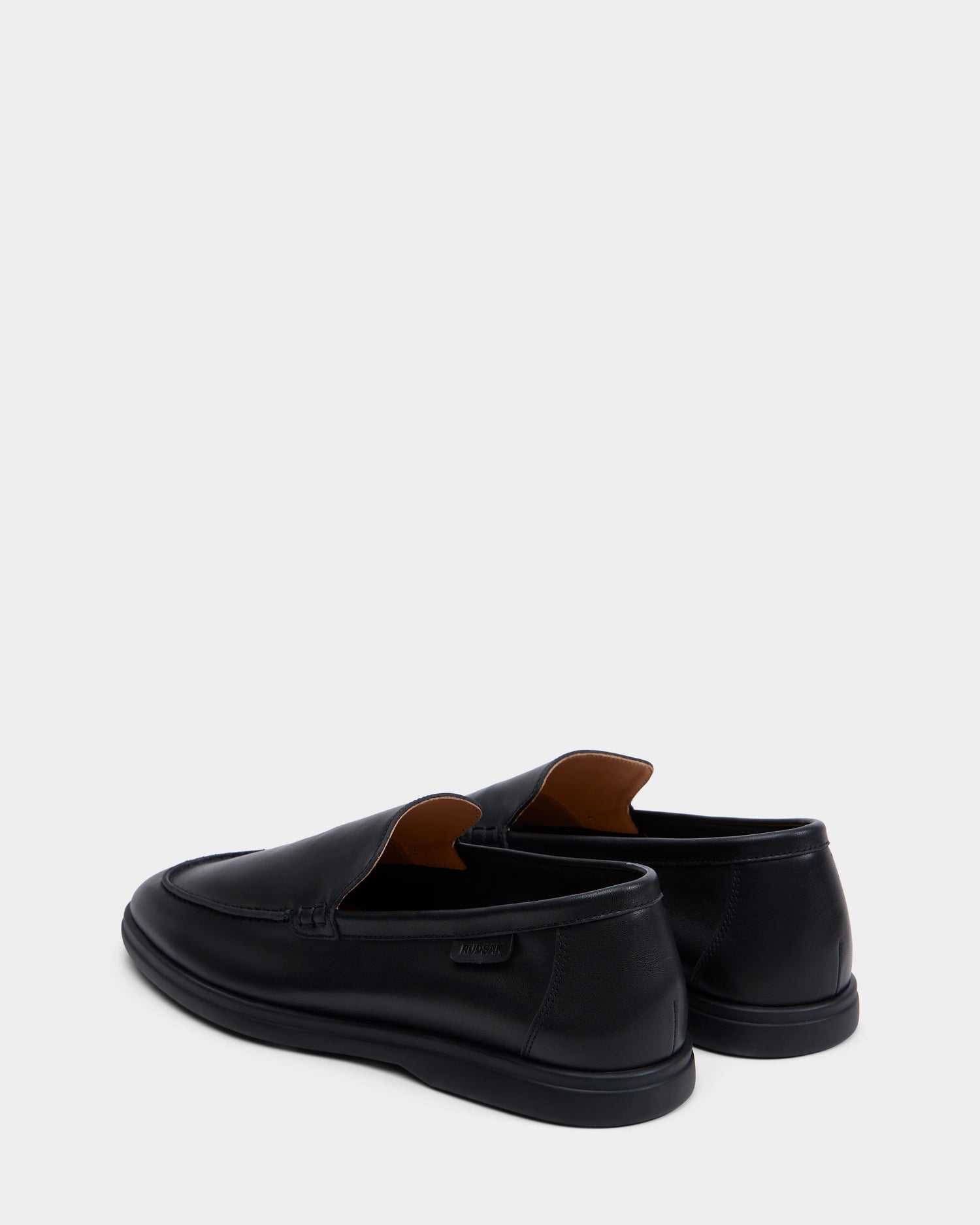The CHANNING - 6225005 Black slip-on loafers by Rudsak are premium men’s shoes, shown on a white background and angled to highlight the back and side details of both black leather shoes.