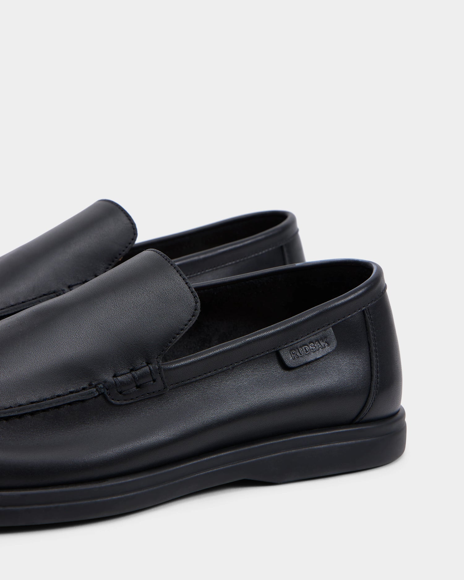 A close-up of the CHANNING - 6225005 Black men’s leather slip-on loafer by Rudsak, featuring subtle stitched detail and a small rectangular logo on the side, photographed on a light gray background—ideal for premium footwear fans.