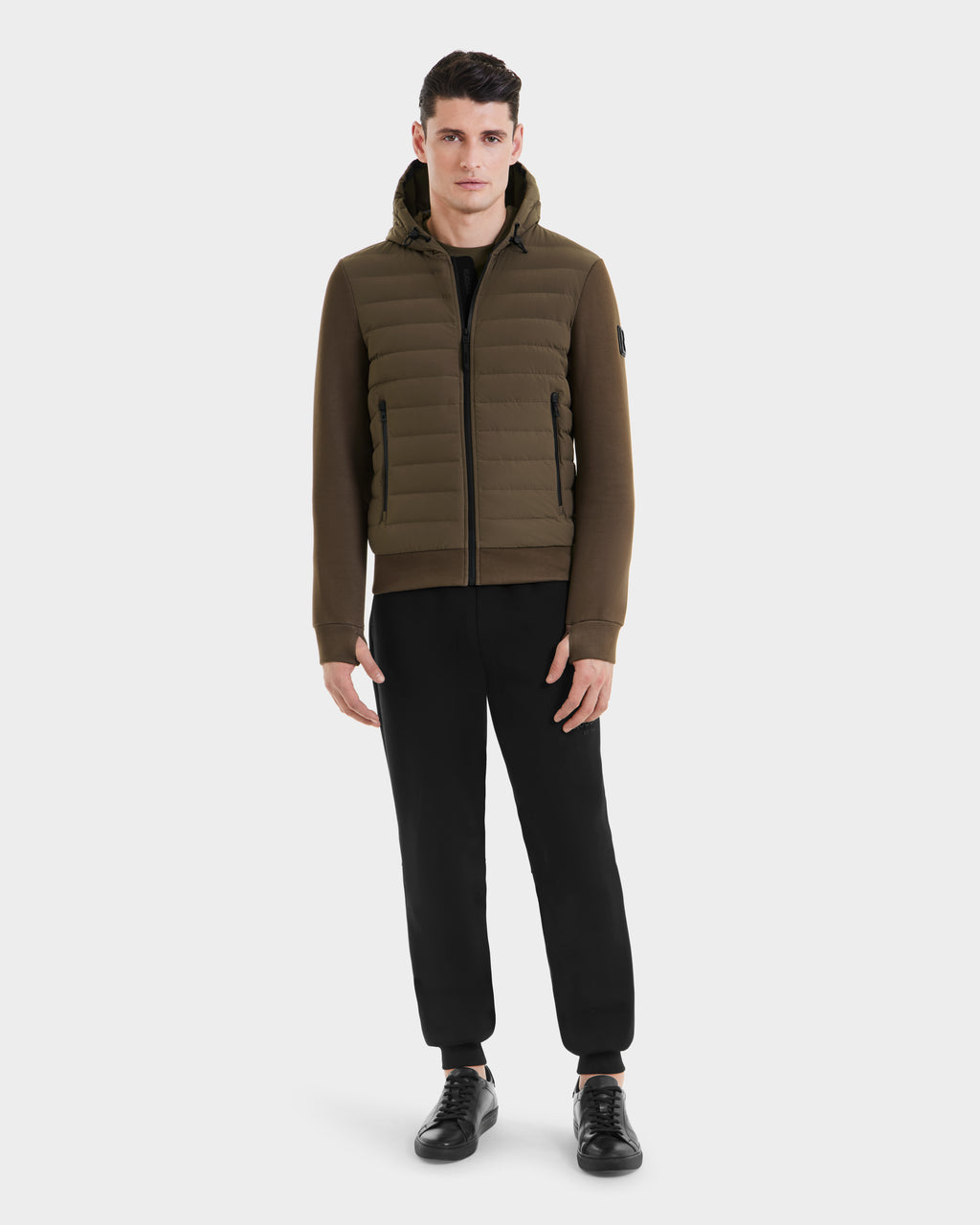 A man faces forward wearing the Rudsak DEE MAT - 6124028 Army mens lightweight hybrid down jacket in brown, paired with black pants and sneakers, against a plain white background.