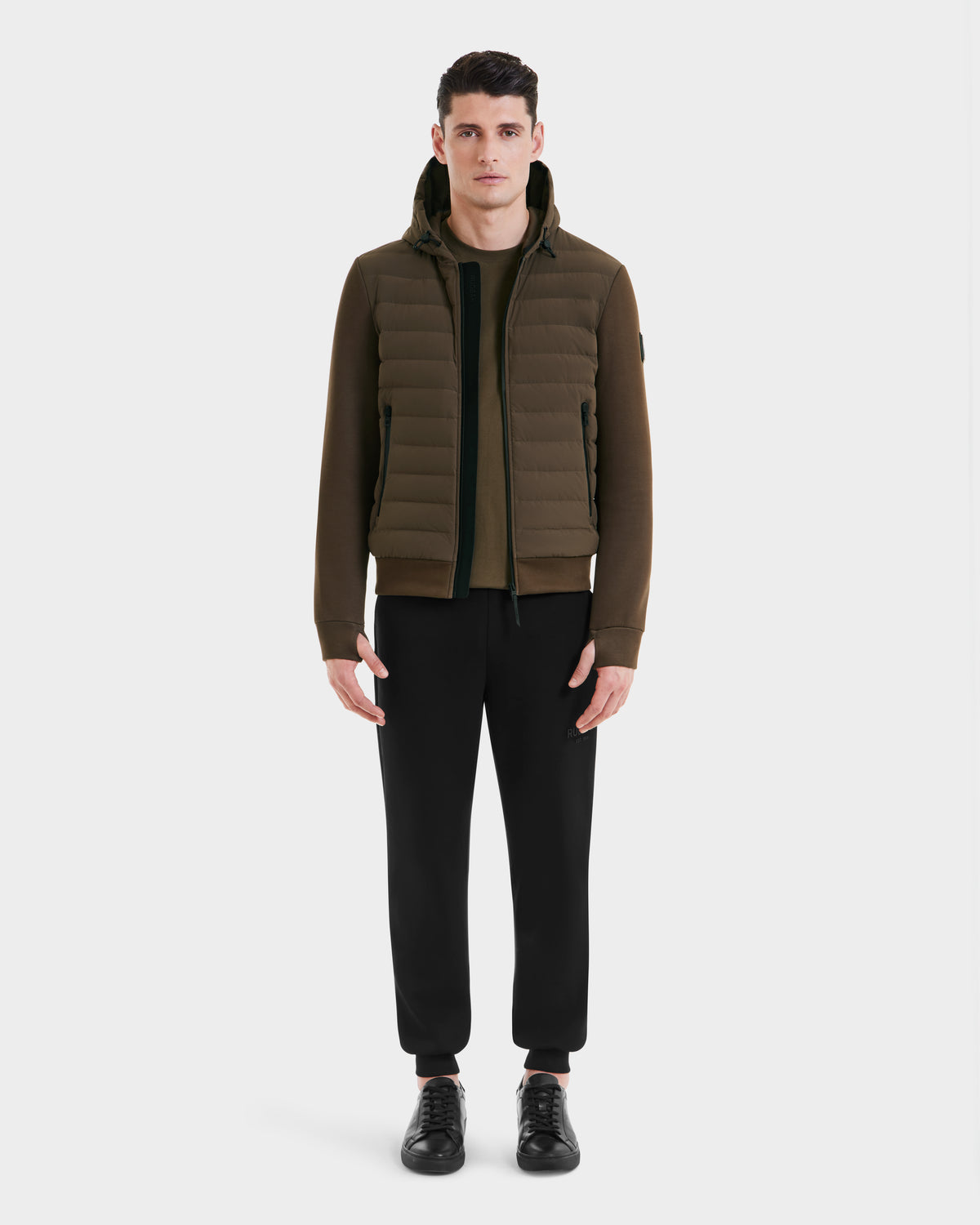 A man faces forward wearing the Rudsak DEE MAT - 6124028 Army mens lightweight hybrid down jacket with a dark green zip-up sweater, black drawstring pants, and black sneakers against a plain white background.