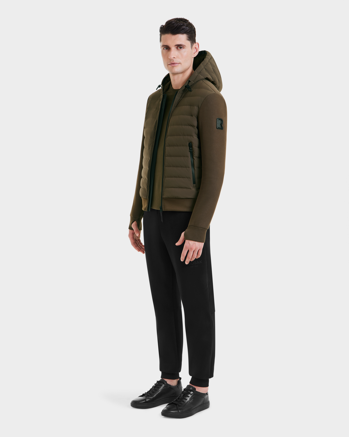 A man faces forward, wearing the Rudsak DEE MAT - 6124028 Army mens lightweight hybrid down jacket in quilted olive-green with a hood, paired with black pants and sneakers, against a plain white background.