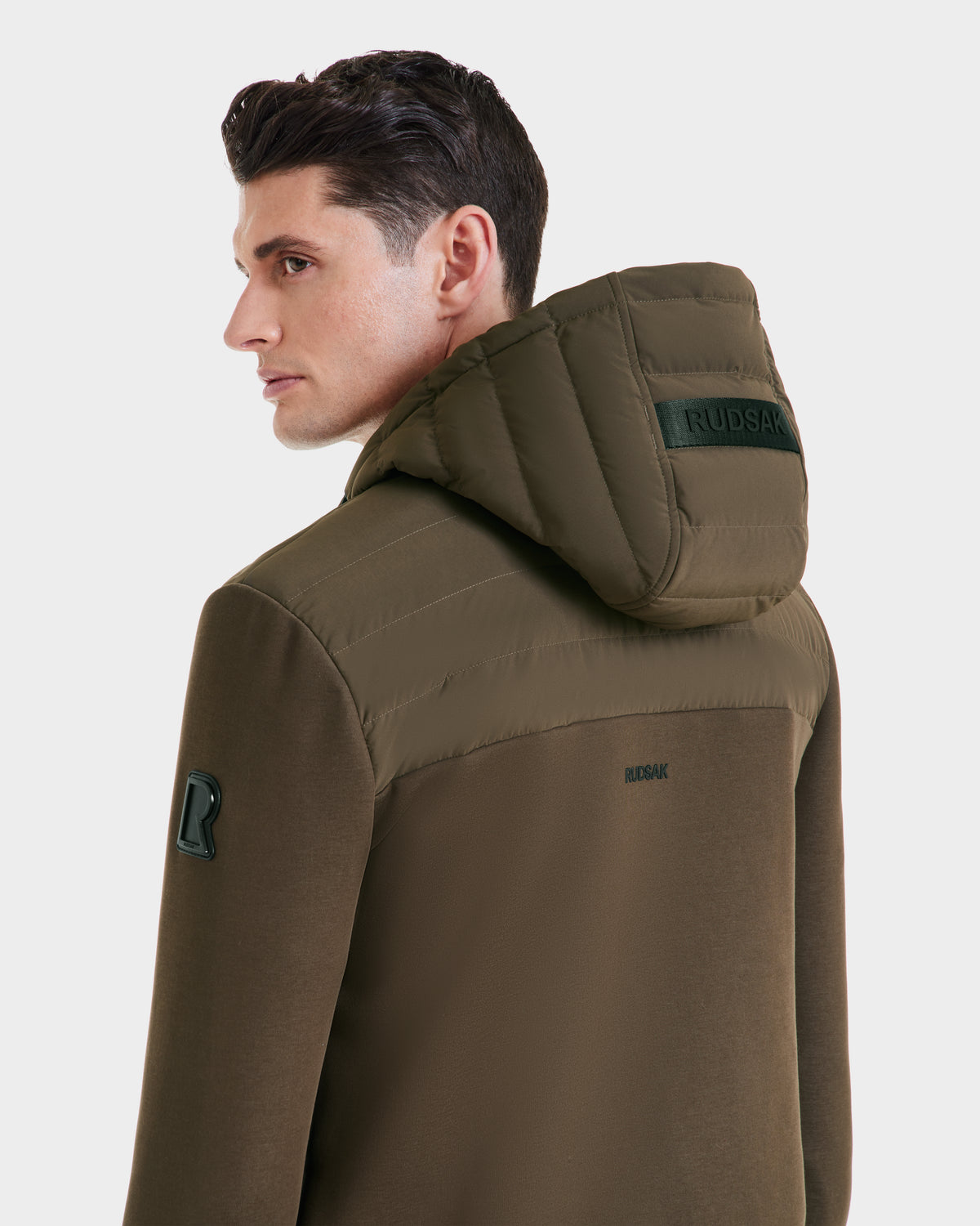 A man with short dark hair is seen from behind wearing the Rudsak DEE MAT - 6124028 Army jacket, an olive green lightweight hybrid down style with a hood. The Rudsak brand name appears on the back of both the hood and jacket.