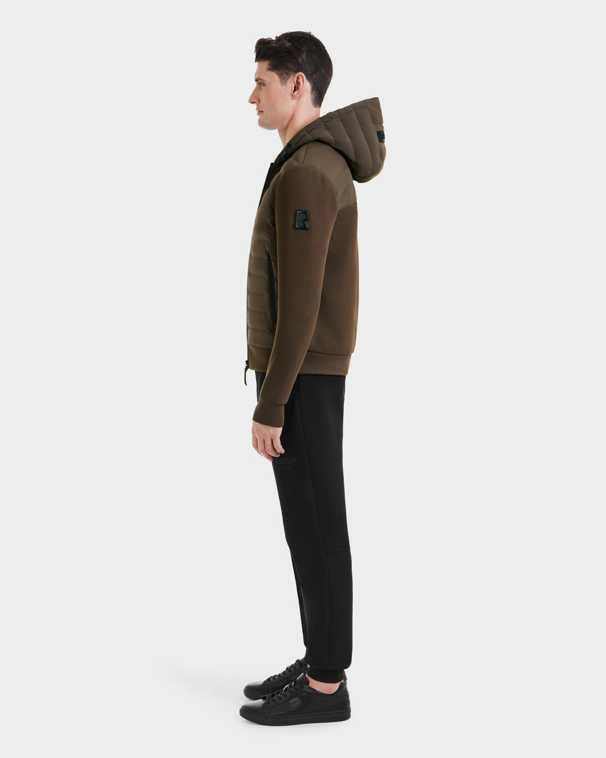 A man stands in profile against a white background, wearing the Rudsak DEE MAT - 6124028 Army mens lightweight hybrid down jacket with quilted detailing, paired with black pants and black sneakers.