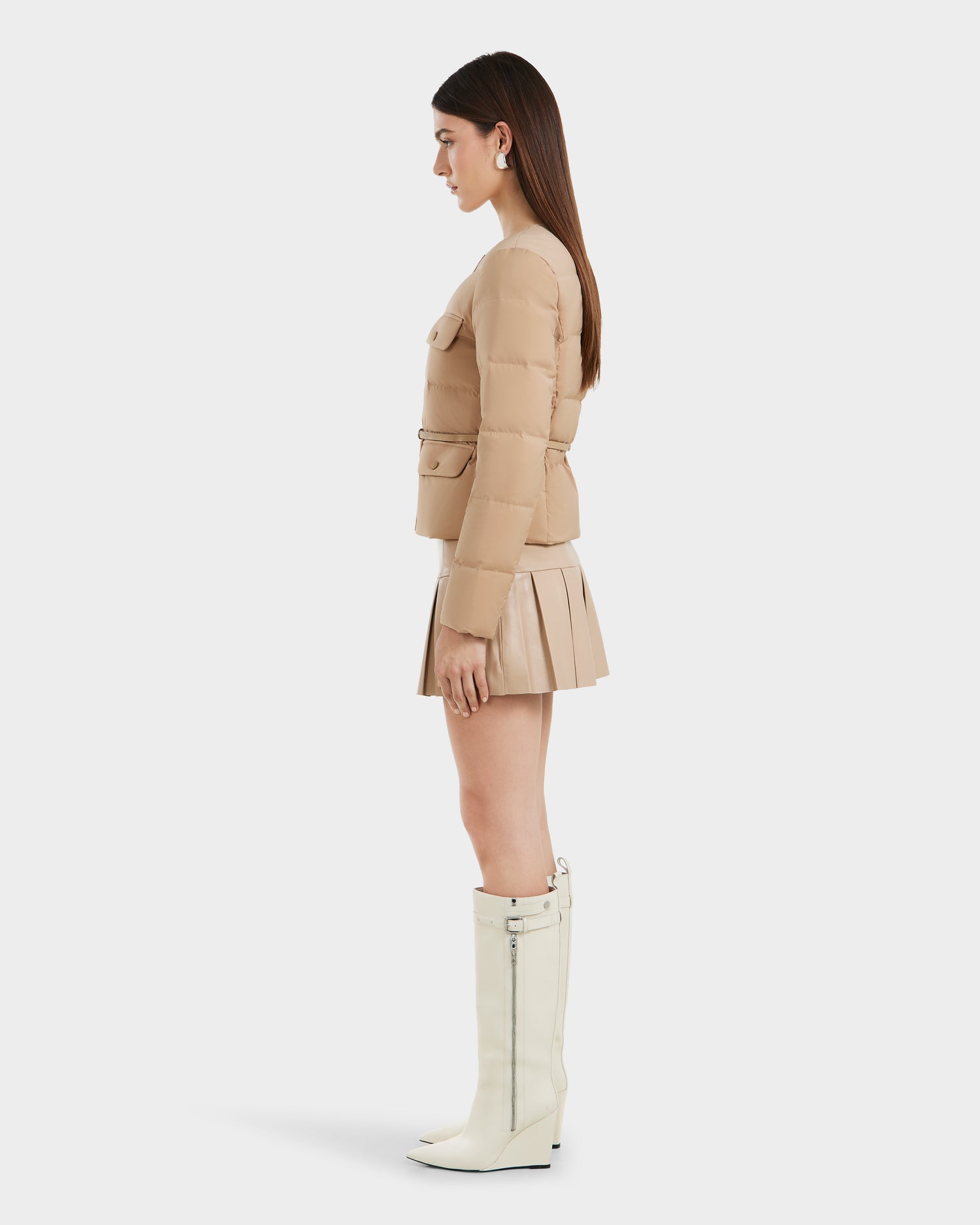 A woman in profile wears the Rudsak FRANCESCA - 8125021 Almond quilted puffer jacket, paired with a matching pleated mini skirt and knee-high white zip boots, set against a plain white background.