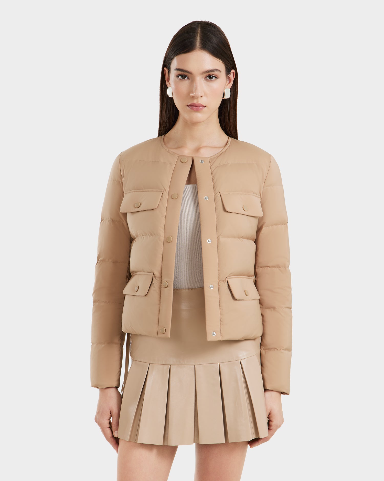 A woman with long dark hair models the Rudsak FRANCESCA - 8125021 Almond puffer jacket, featuring snap buttons and flap pockets, styled over a light top and beige pleated skirt with a vegan leather belt, against a plain white background.