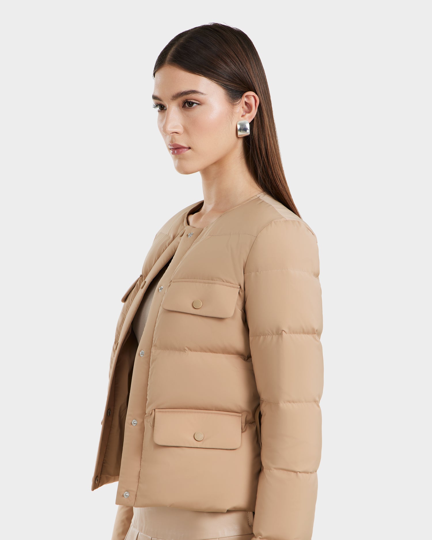 A woman with long, straight brown hair wears the Rudsak FRANCESCA - 8125021 Almond puffer jacket with front pockets and large silver hoop earrings, posing against a plain white background.