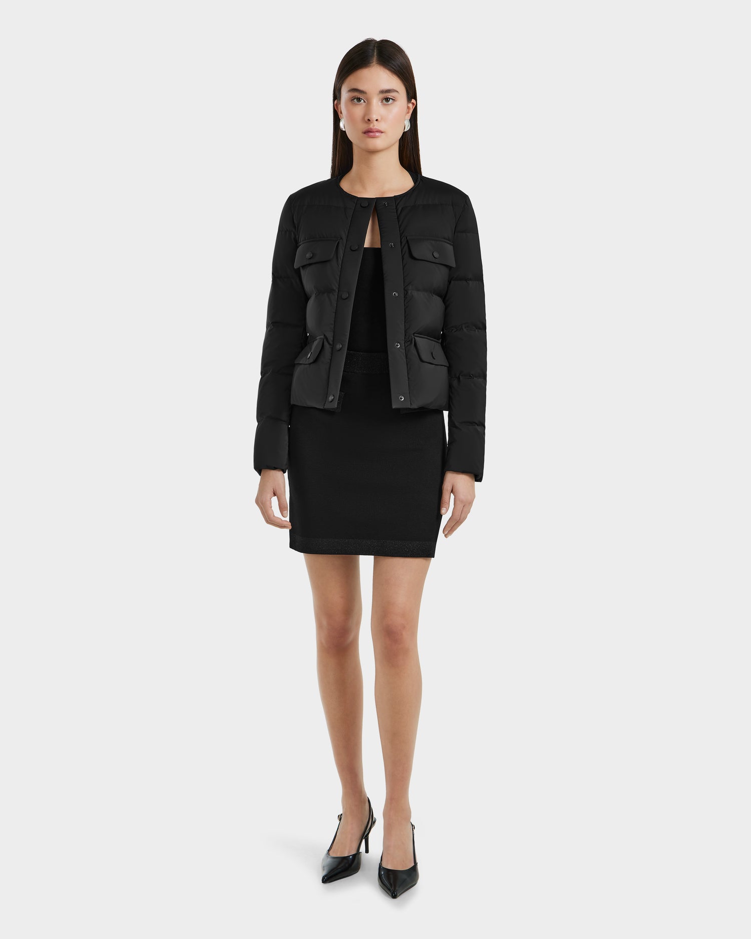 A woman faces forward against a white background wearing the Rudsak FRANCESCA - 8125021 Black quilted puffer jacket over a black outfit, cinched with a vegan leather belt, and paired with black pointed-toe heels.