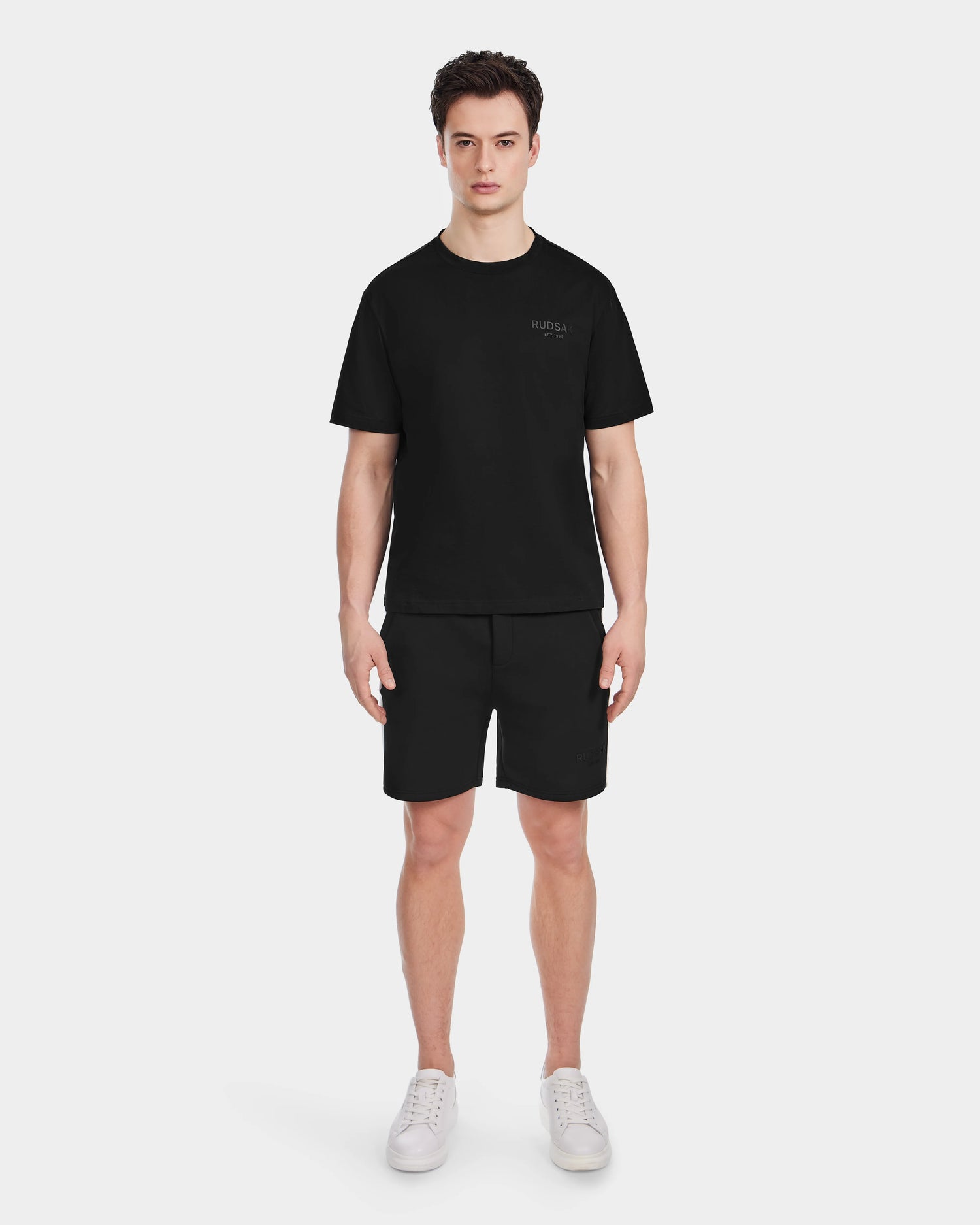 A person stands facing forward, wearing the Rudsak GRANT - 6525036 Black shorts, styled with a mens premium cotton crewneck tee and white sneakers against a plain white background.