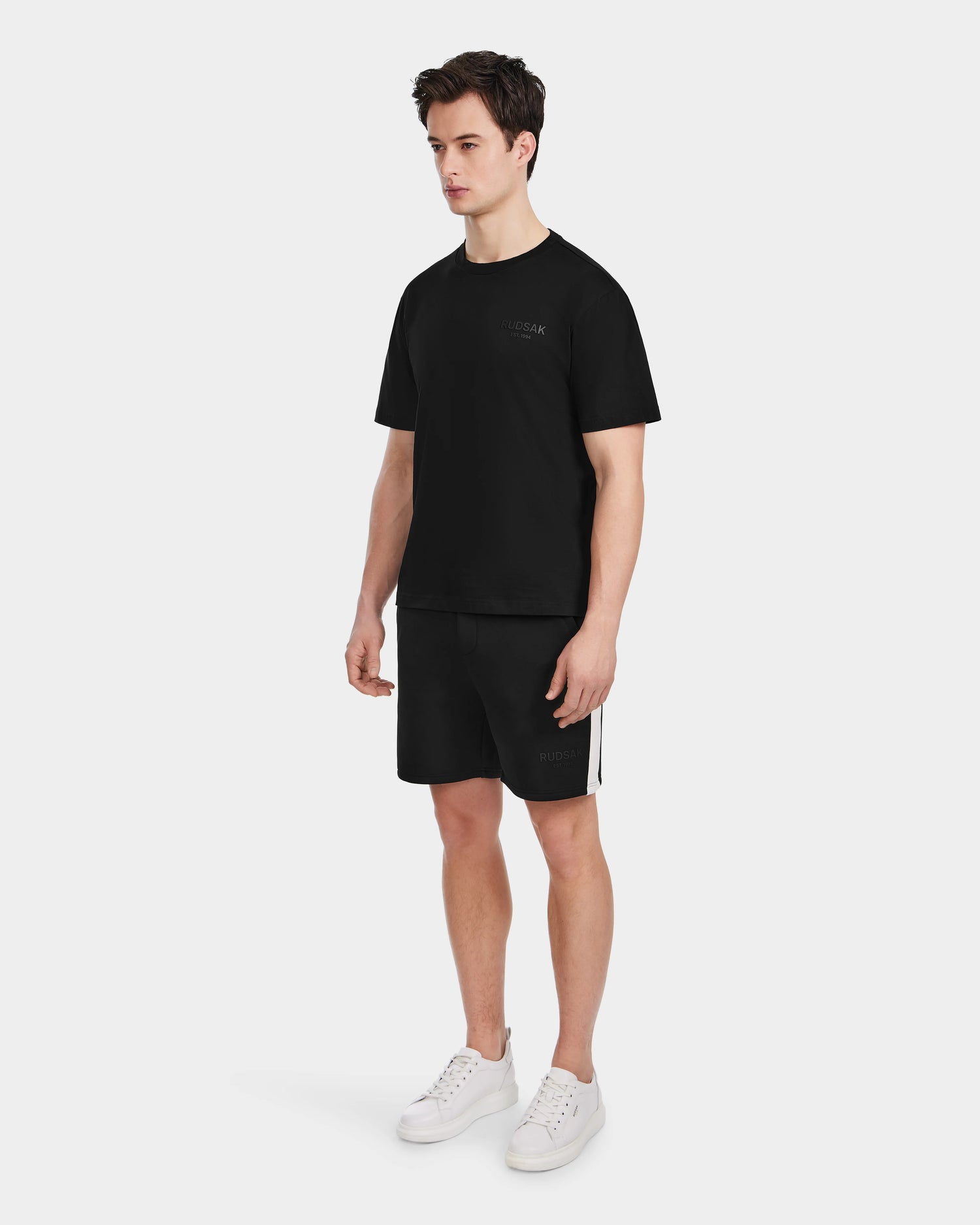 A young man stands against a white background in the Rudsak GRANT - 6525036 Black premium cotton mens tee, paired with black shorts and white sneakers. His look is simple and casual, and he gazes slightly to his left.