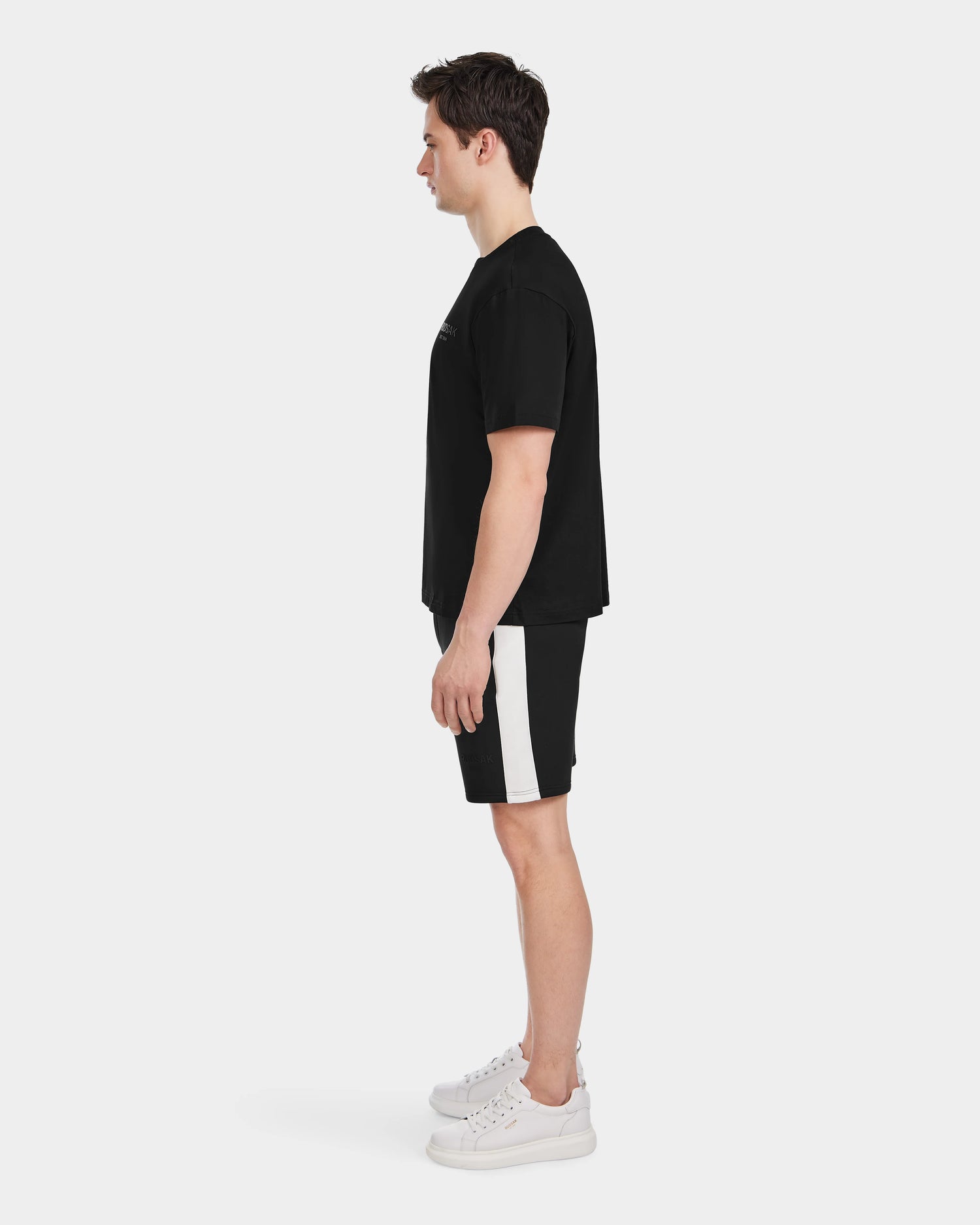 A man stands in profile against a plain white background, wearing the Rudsak GRANT - 6525036 Black shorts with a white side stripe, paired with a premium cotton mens tee and white sneakers.