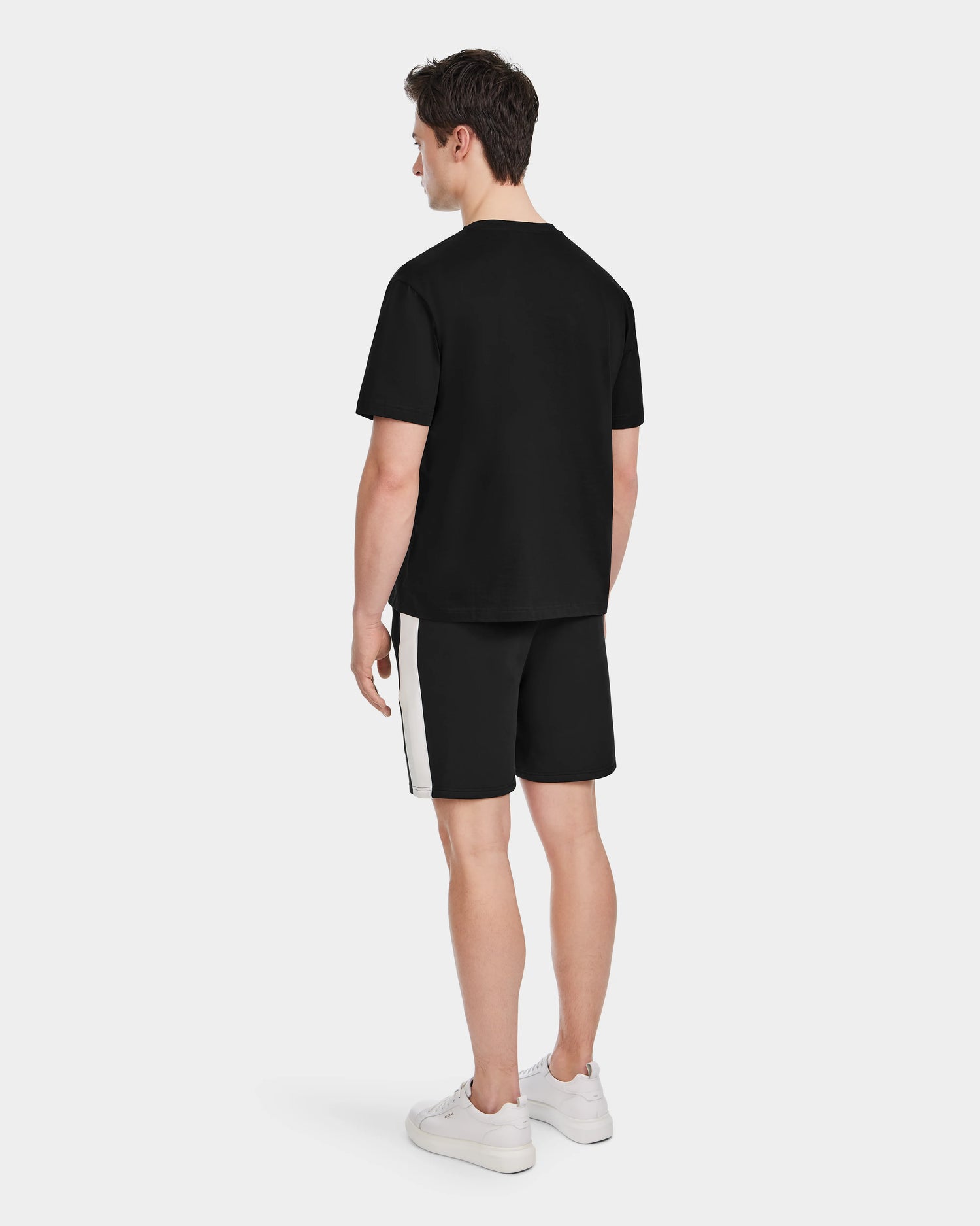 A man faces away, wearing the Rudsak GRANT - 6525036 Black premium cotton mens crewneck tee with black shorts featuring a white side stripe and white sneakers against a plain light background.