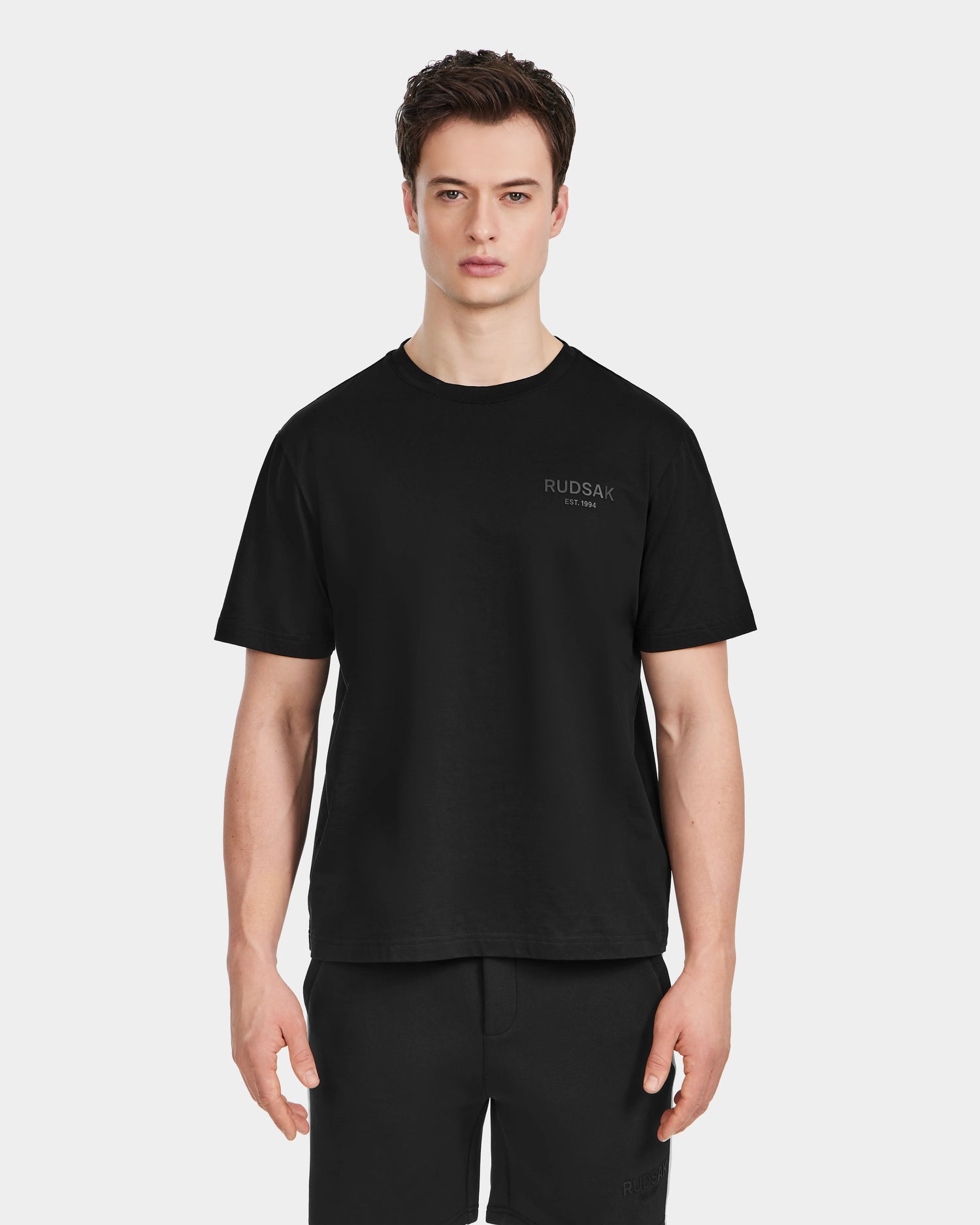 A young man faces forward wearing the Rudsak GRANT - 6525036 Black premium cotton crewneck tee with RUDSAK EST. 1994 in small print on the upper left chest, paired with black shorts against a plain white background.