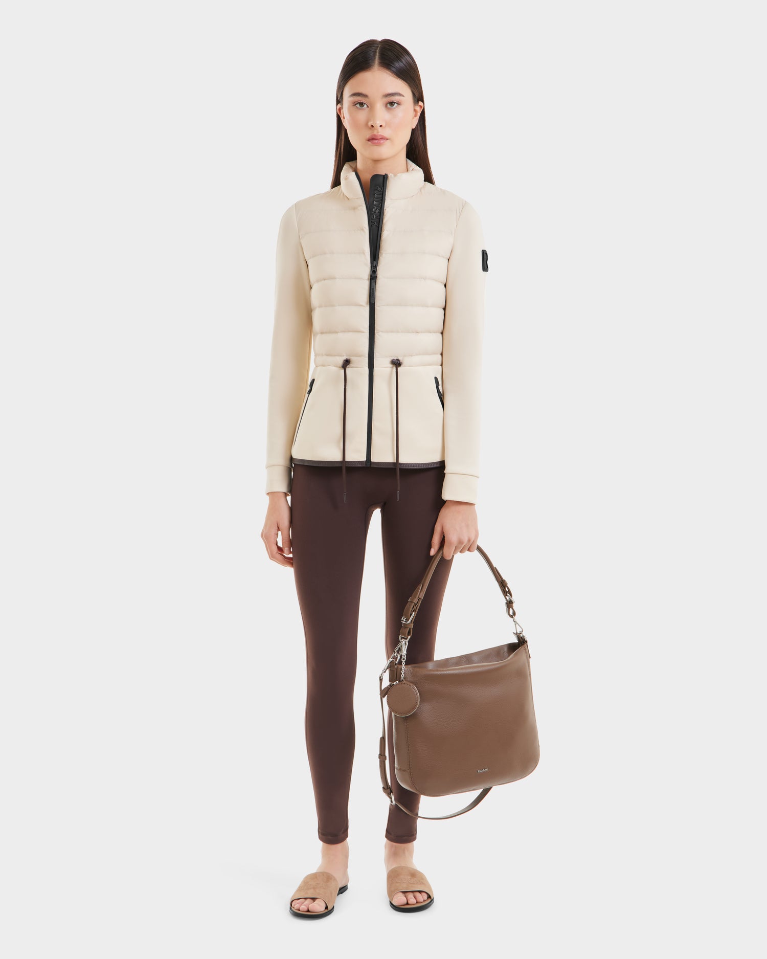 A woman with straight dark hair and a neutral expression stands facing forward in a Rudsak JOSEPHINE - L125011 Vanilla hybrid jacket, brown leggings, beige slide sandals, and holds a large brown shoulder bag. The background is white.