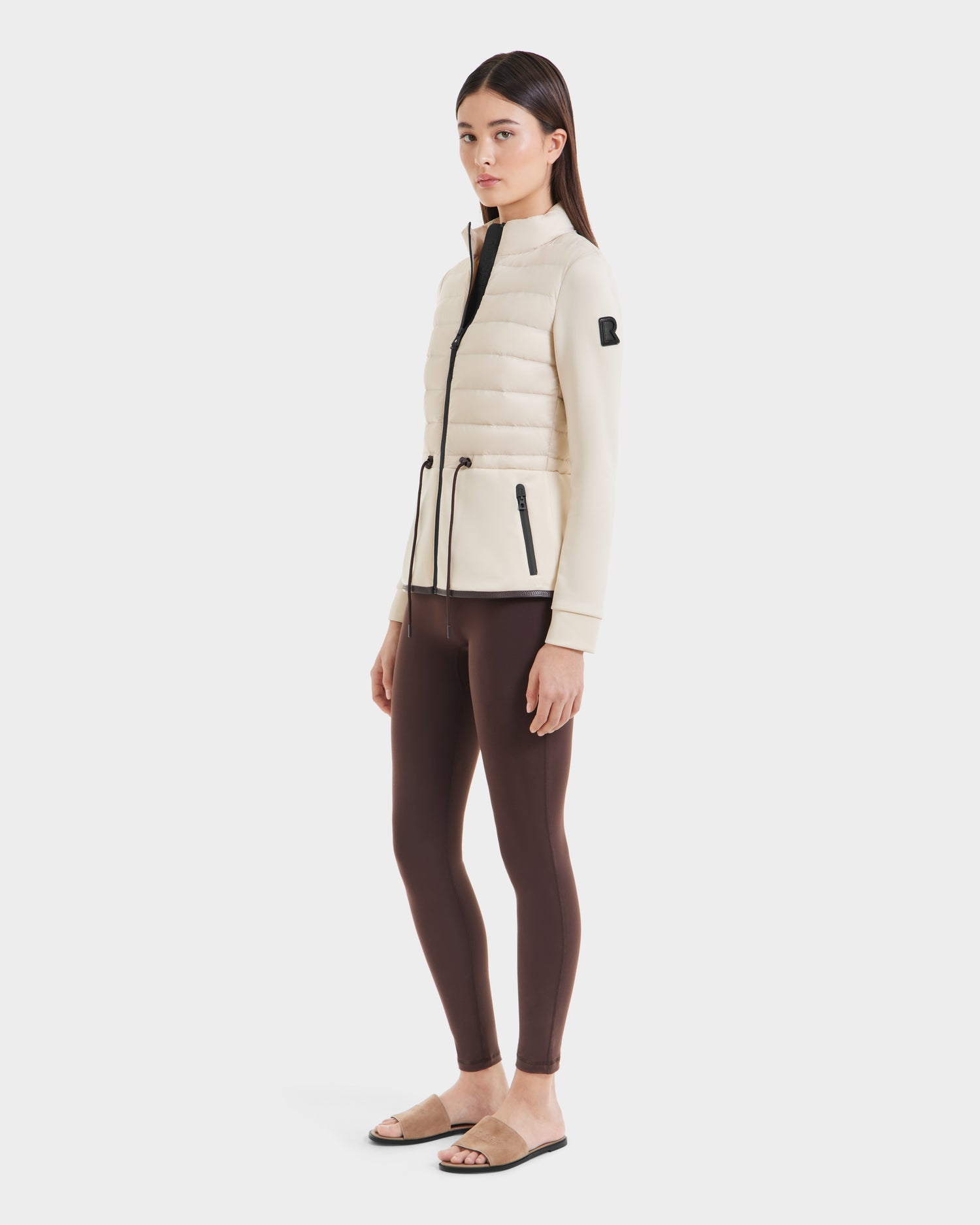A woman stands facing slightly left, wearing the Rudsak JOSEPHINE - L125011 Vanilla cream hybrid jacket with zipper pockets, brown leggings, and beige slide sandals against a plain white background.