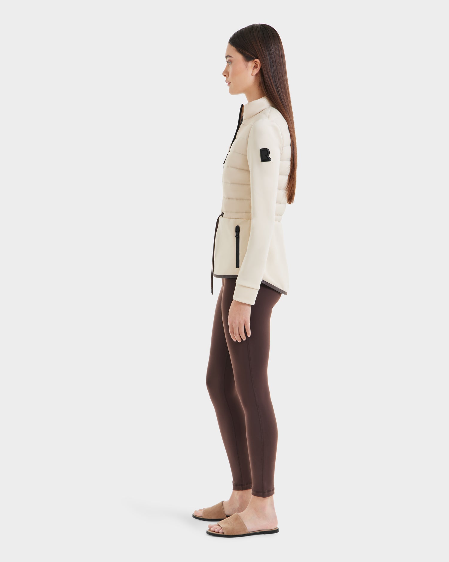 A woman in profile against a white backdrop wears the Rudsak JOSEPHINE - L125011 Vanilla hybrid jacket with brown leggings and tan sandals. Her long straight hair is down, and her arms rest naturally at her sides.