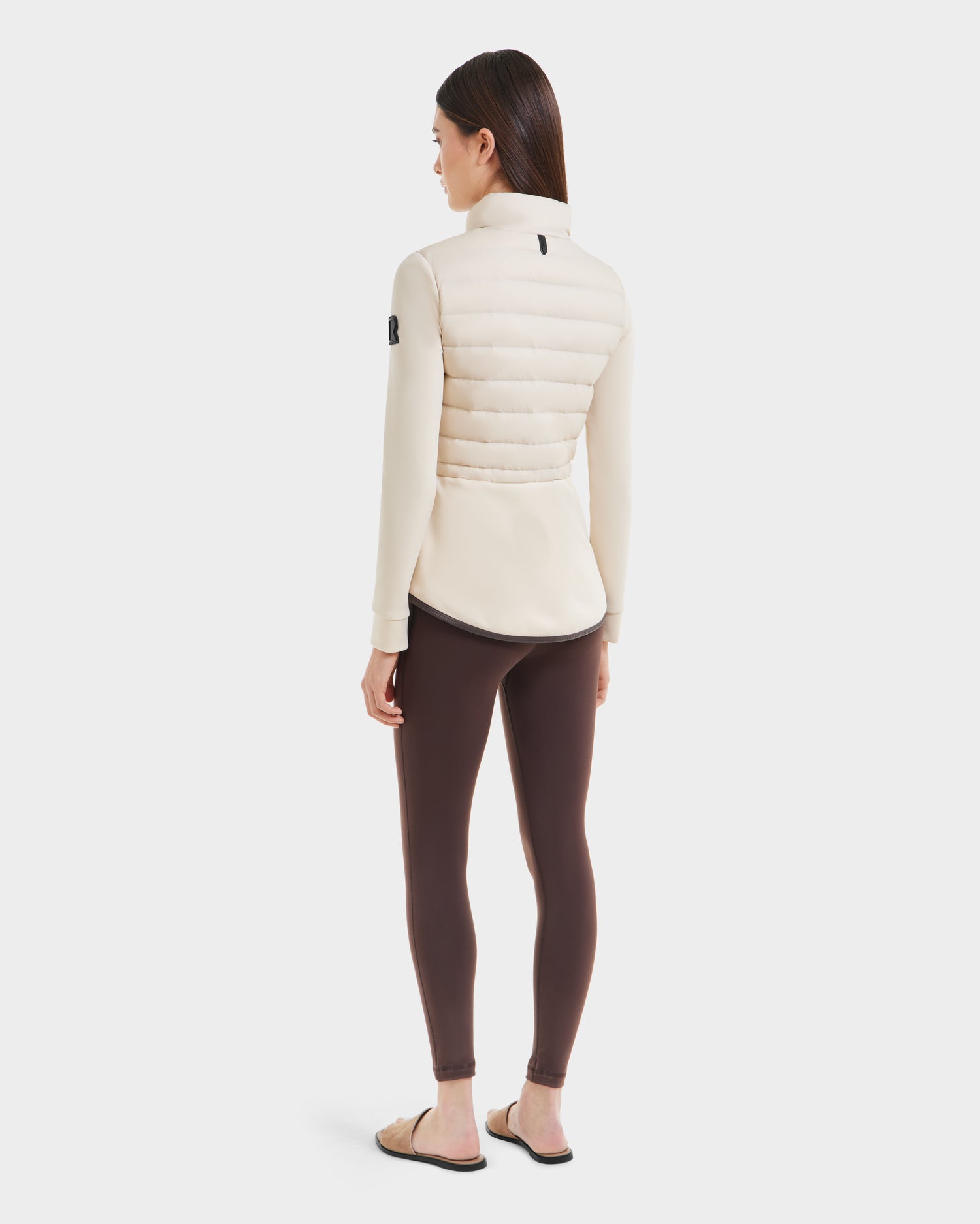 A woman with straight brown hair faces away, wearing the Rudsak JOSEPHINE - L125011 Vanilla quilted hybrid jacket, styled with brown leggings and beige slide sandals against a plain white background.