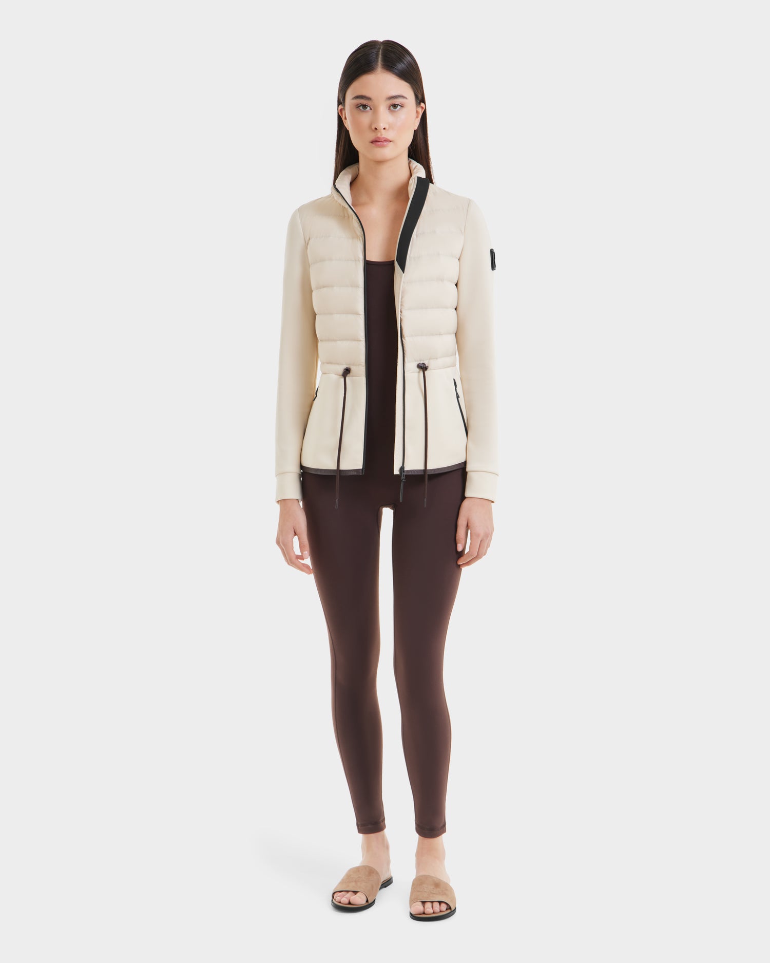 A woman stands facing forward against a white background, wearing the Rudsak JOSEPHINE - L125011 Vanilla jacket with black trim, brown leggings, and beige slide sandals.