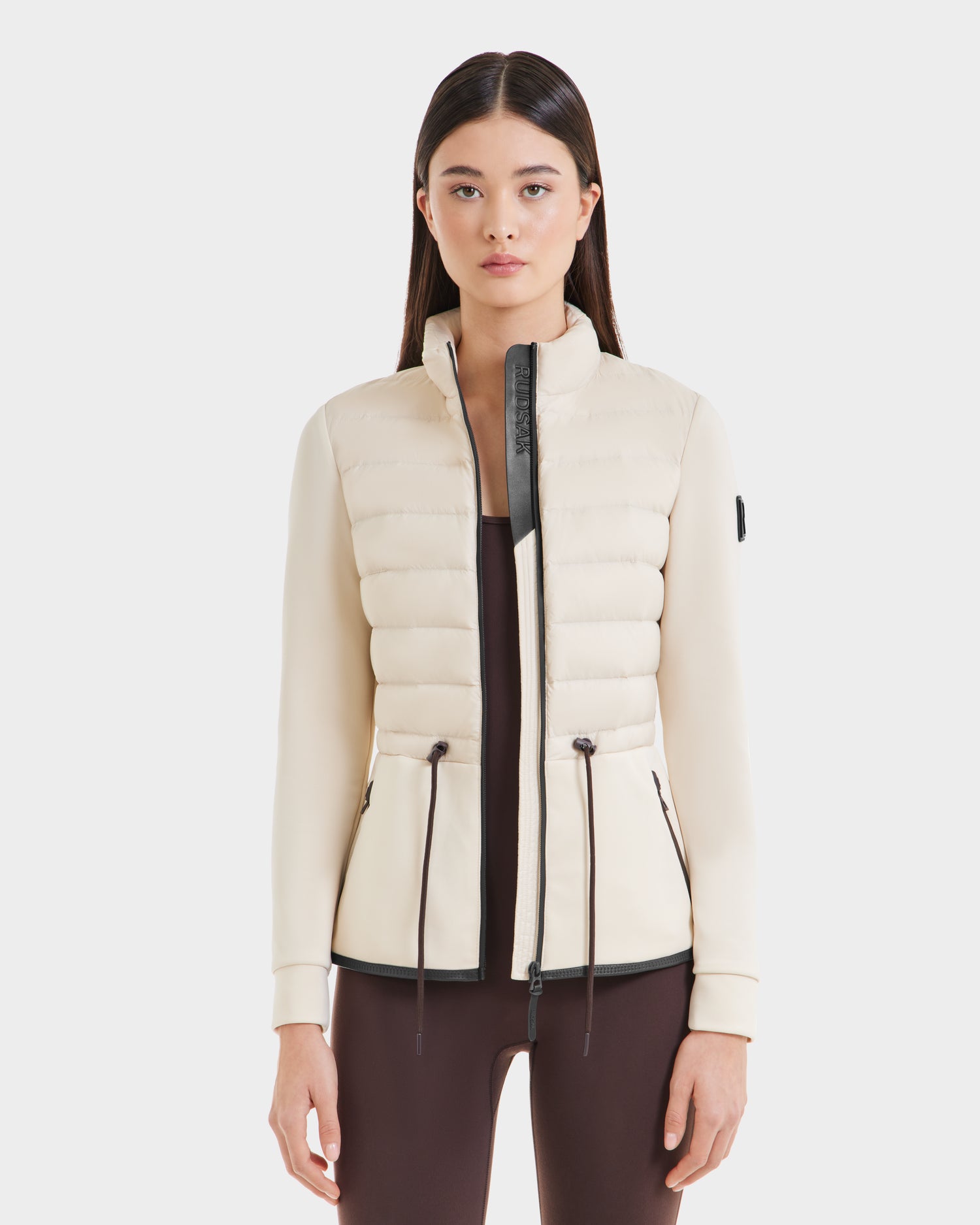 A woman with straight, dark hair models the Rudsak JOSEPHINE - L125011 Vanilla hybrid jacket in cream recycled nylon with quilted panels and waist drawstrings, paired with dark pants against a plain white background.