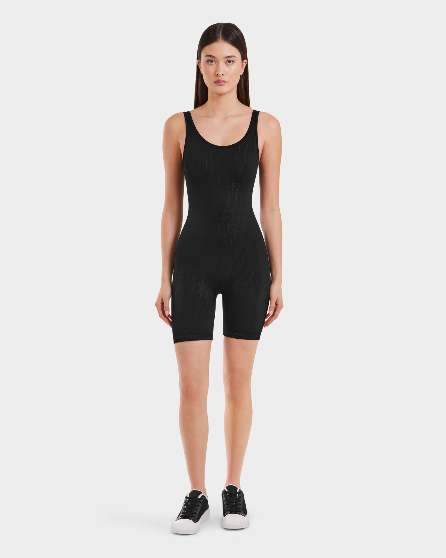 A woman with straight brown hair faces forward, wearing the KAILEY - 8523012 Black Pattern romper by Rudsak and black sneakers with white soles against a plain white background.