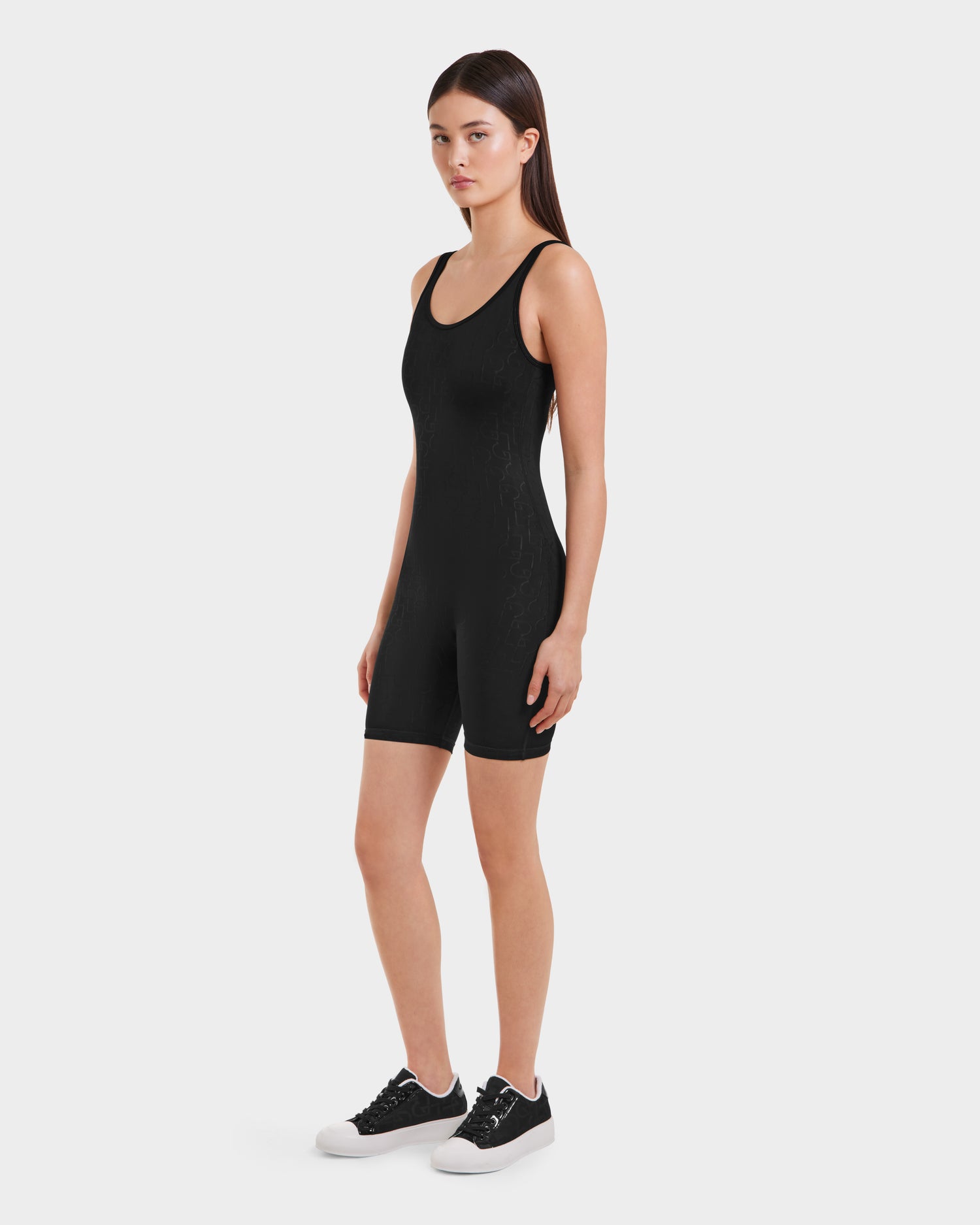 A woman with straight brown hair and a neutral expression stands against a white background, wearing the Rudsak KAILEY - 8523012 Black Pattern fitted sleeveless romper with black sneakers. Her arms are relaxed at her sides.