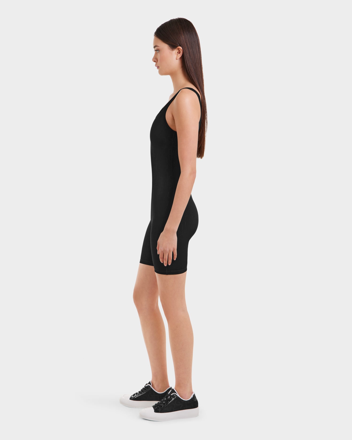 A woman stands in profile against a white background, wearing the Rudsak KAILEY - 8523012 Black Pattern fitted sleeveless romper and black sneakers with white soles.
