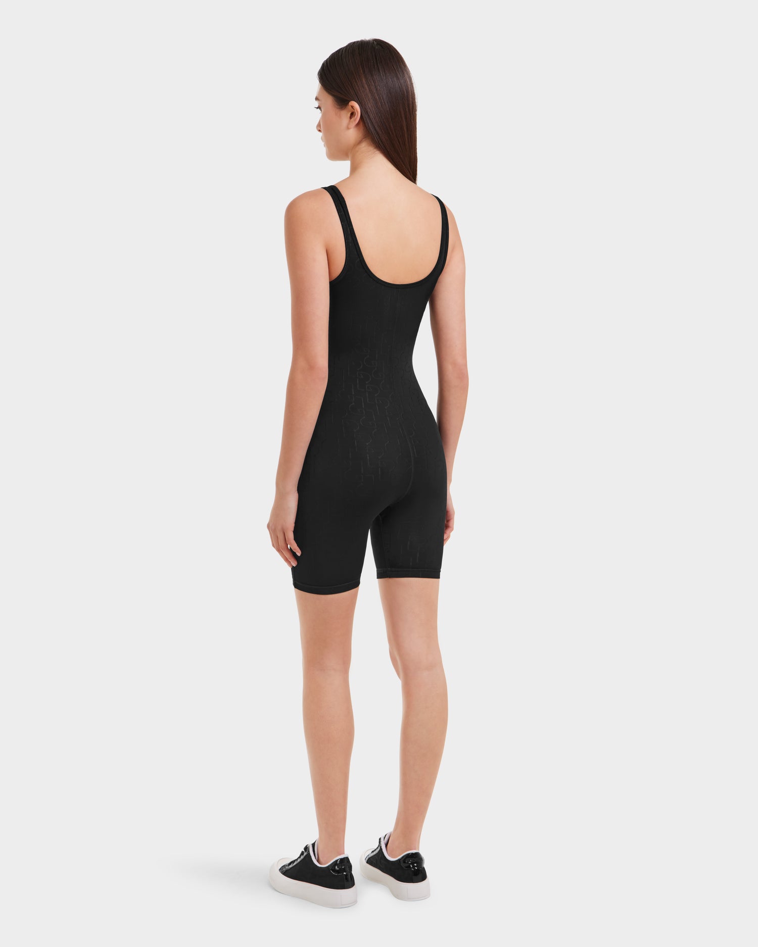 A woman with straight brown hair, seen from behind, wears the Rudsak KAILEY - 8523012 Black Pattern sleeveless romper and black sneakers with white soles against a plain white background.