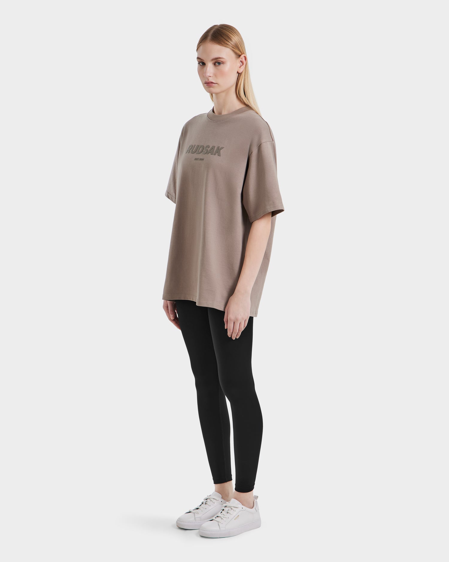 A woman with straight blonde hair wears the Rudsak KARTER - 9524008 Cement W T-shirt, pairing it with black leggings and white sneakers, standing against a plain white background.