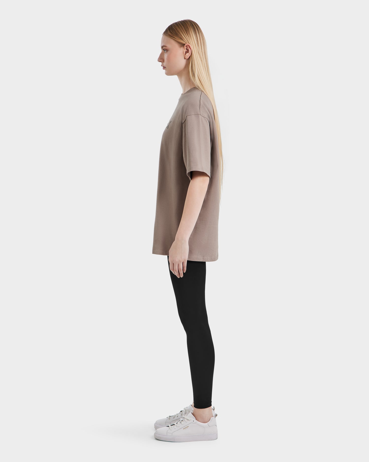 A young woman with long blonde hair stands in profile, wearing the Rudsak KARTER - 9524008 Cement W oversized unisex T-shirt in brown, paired with black leggings and white sneakers against a plain white background.