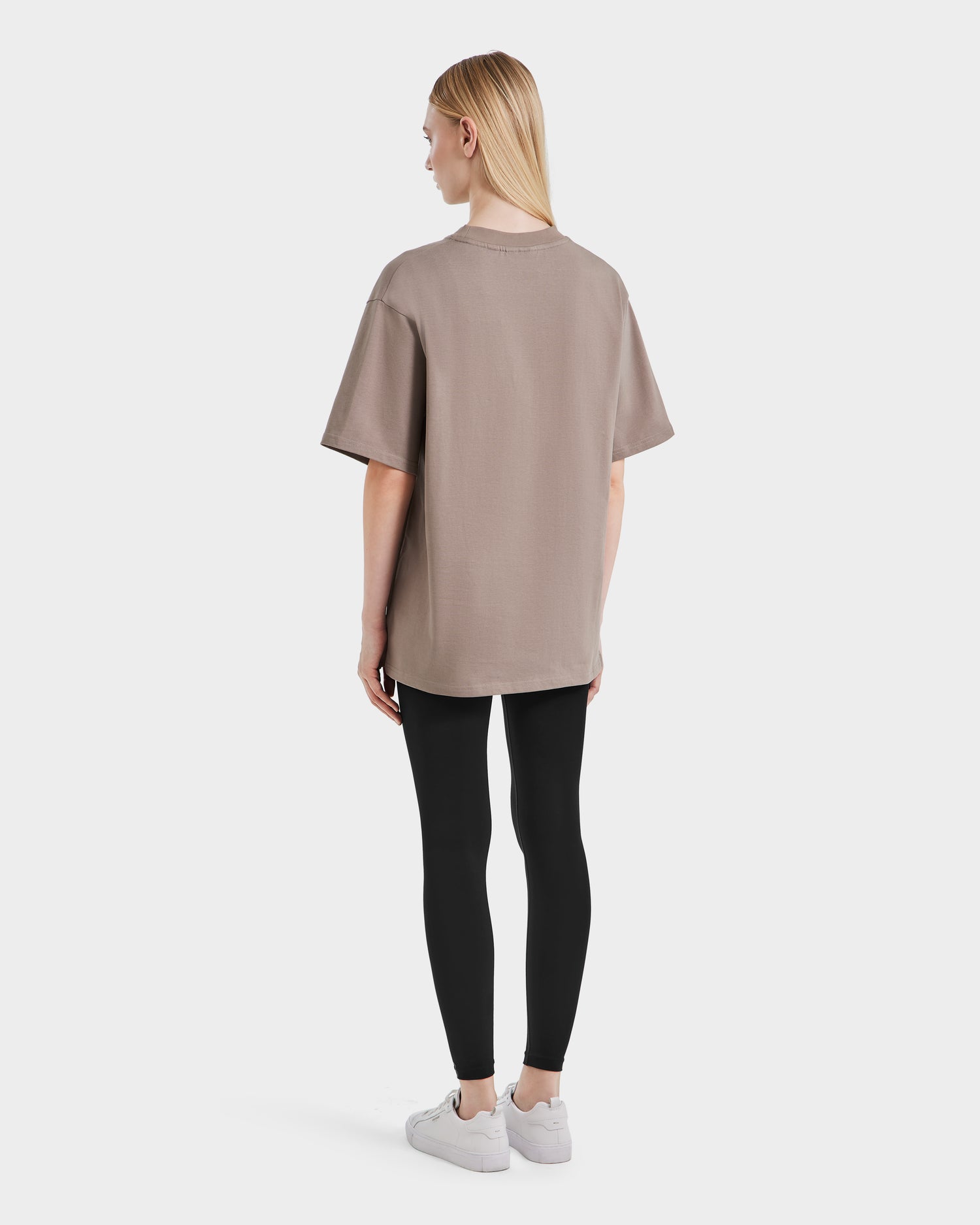 A person with straight blonde hair stands facing away, wearing the Rudsak KARTER - 9524008 Cement W relaxed fit taupe T-shirt in premium cotton, paired with black leggings and white sneakers against a plain white background.