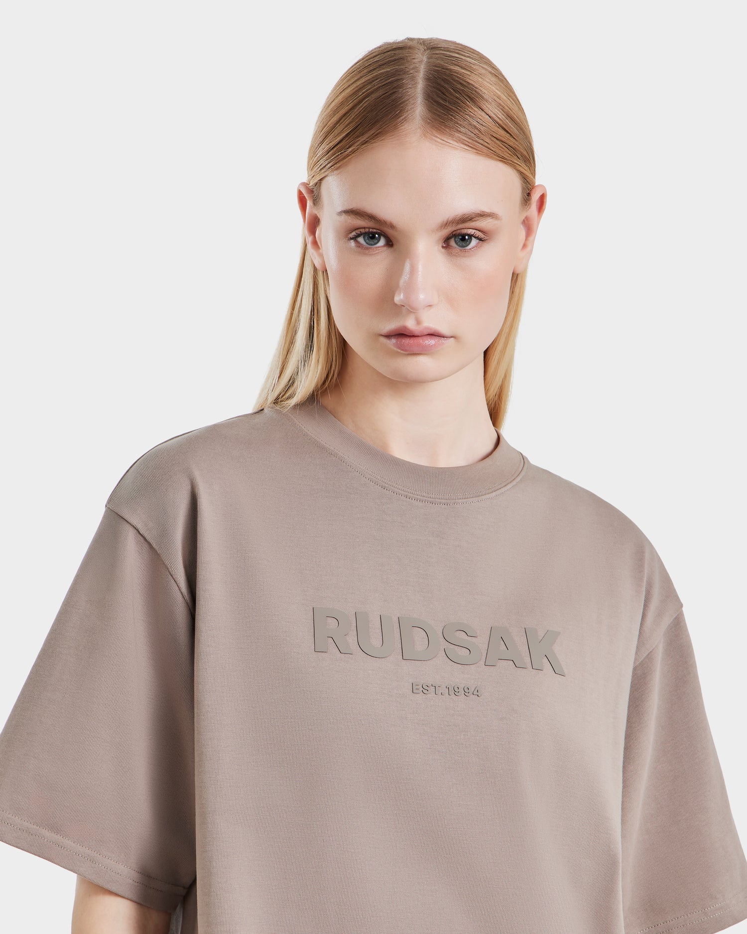 A woman with straight blonde hair wears the Rudsak KARTER - 9524008 Cement W taupe oversized unisex T-shirt, made from premium cotton and featuring "RUDSAK EST. 1994" on the front, standing against a white background.