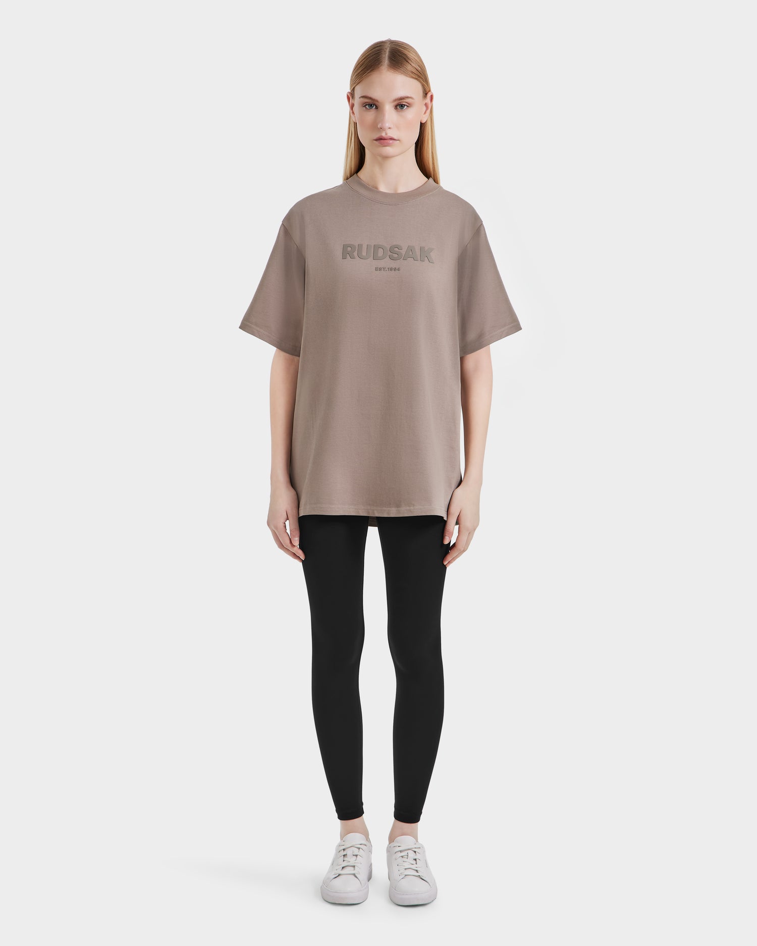 A woman faces forward, wearing a relaxed fit Rudsak KARTER - 9524008 Cement W oversized unisex T-shirt, paired with black leggings and white sneakers, against a plain white background.
