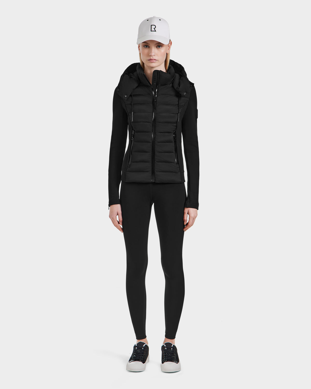 A person faces forward wearing the Rudsak MABS - 8124825 Black womens jacket over black leggings, a white baseball cap, and black sneakers with white soles, all set against a plain white background.